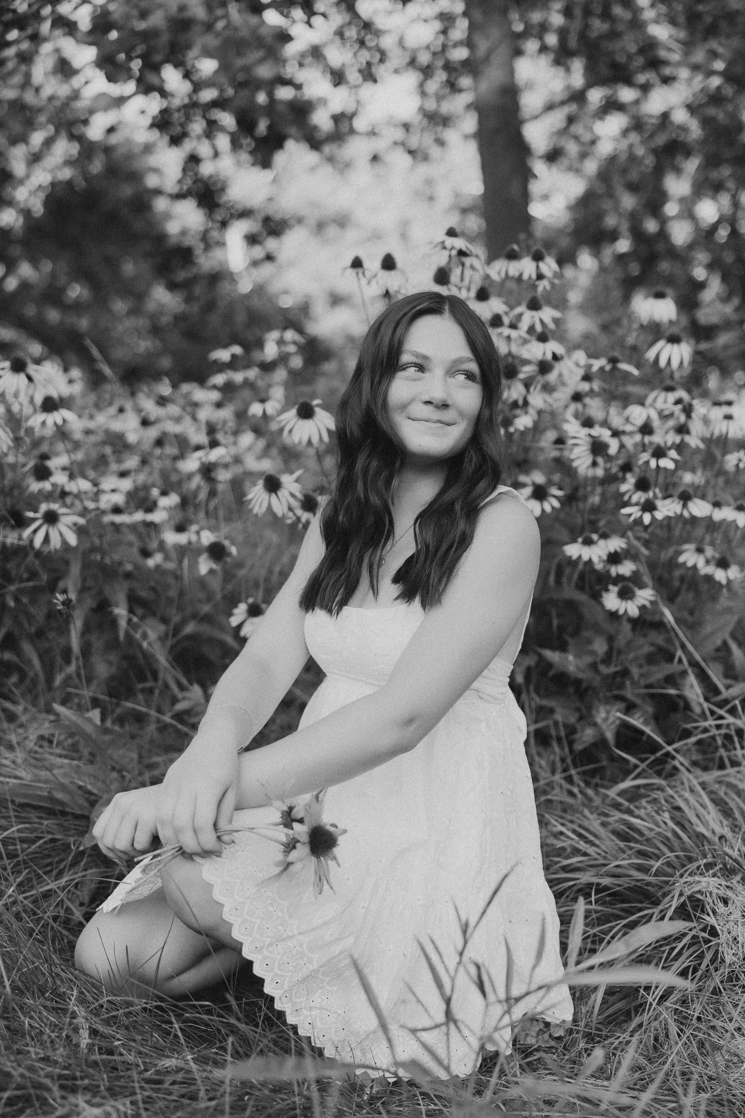  a high school senior kneels in the grass in front of a flower bunch and smiles over her shoulder 