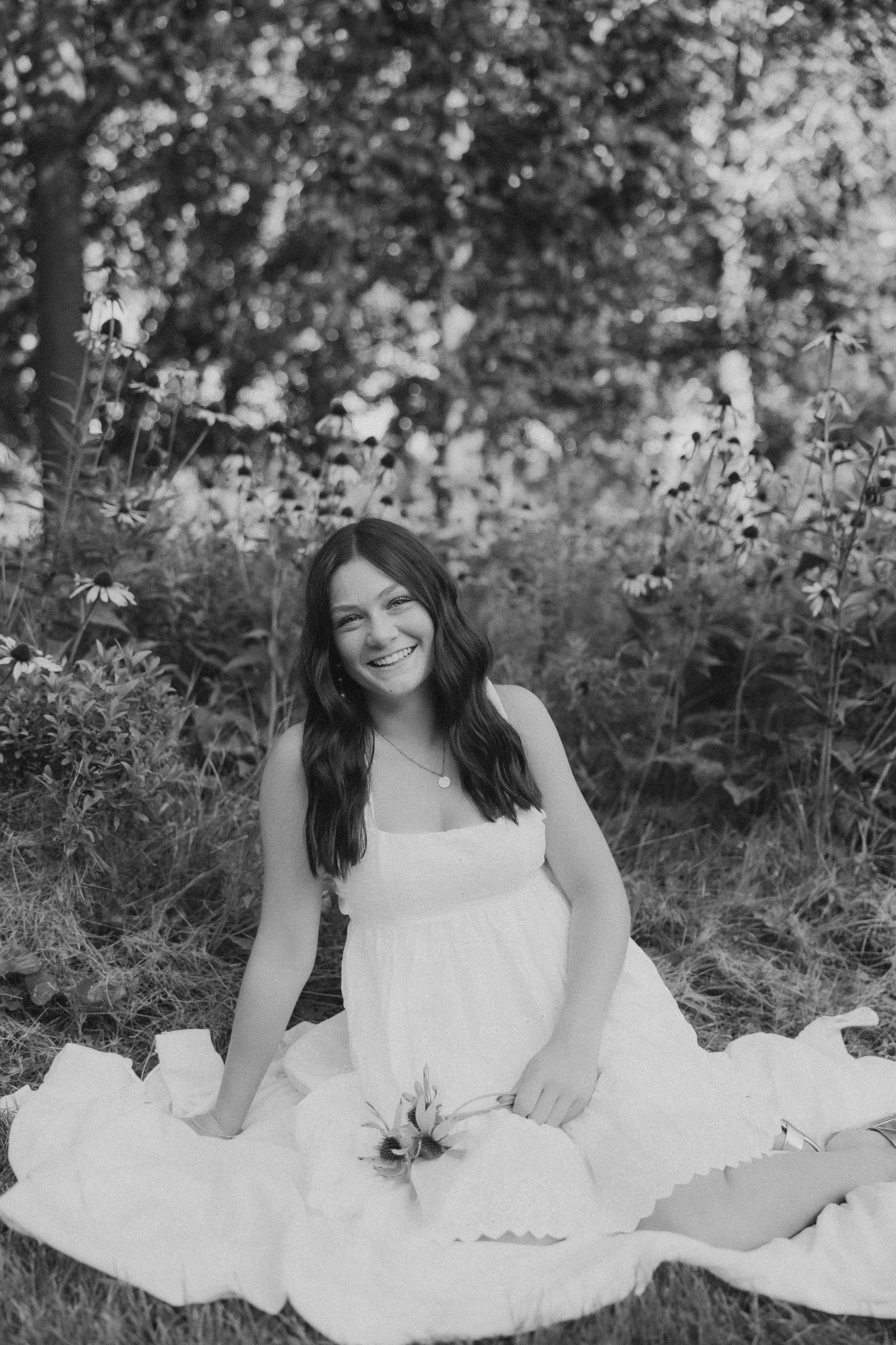 a high school senior sits on a blanket in a white dress in a wild flower field for senior pictures near me 