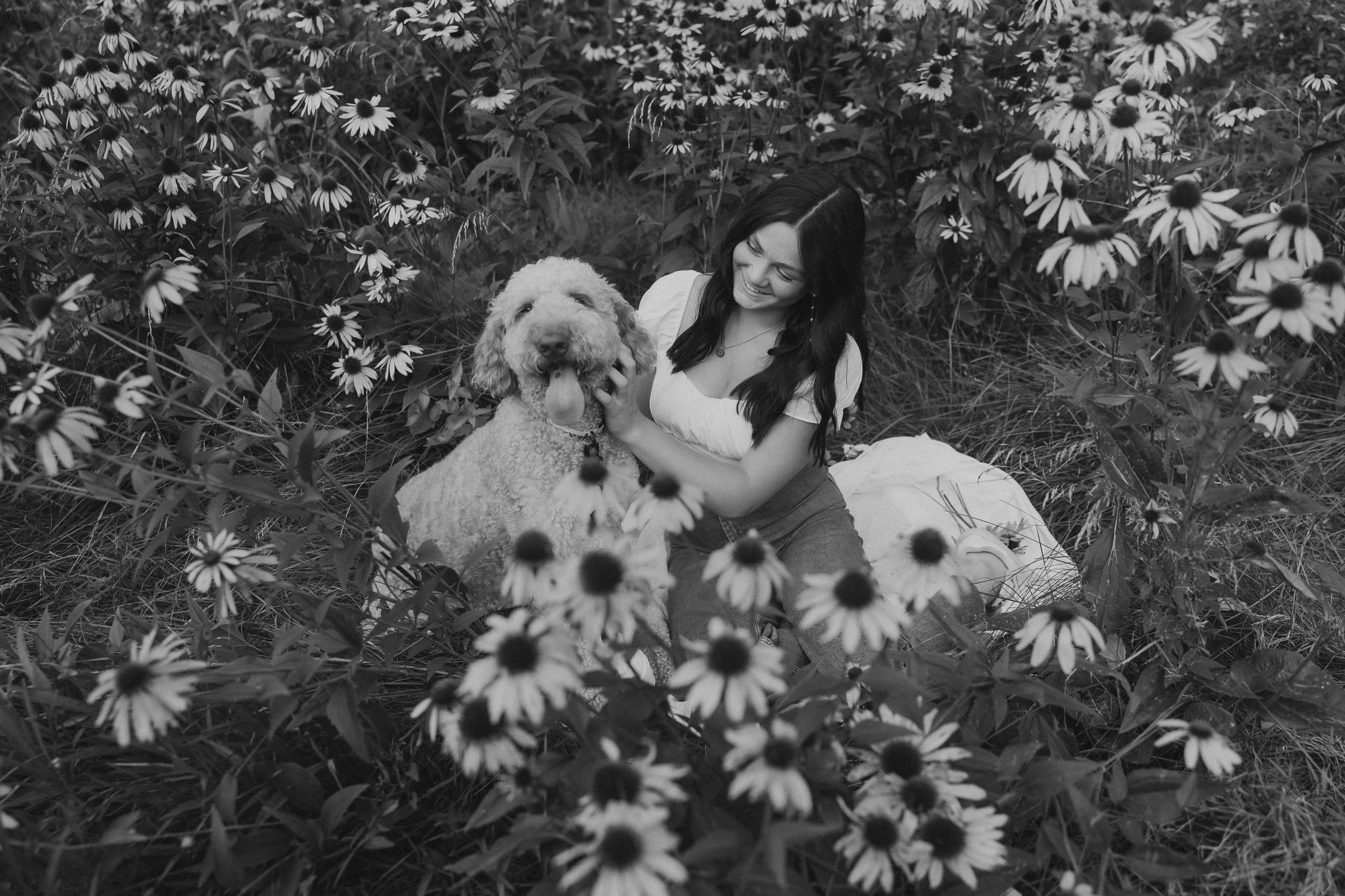  olivia looks over at her dog while petting him in a field of flowers for her senior pictures near me 