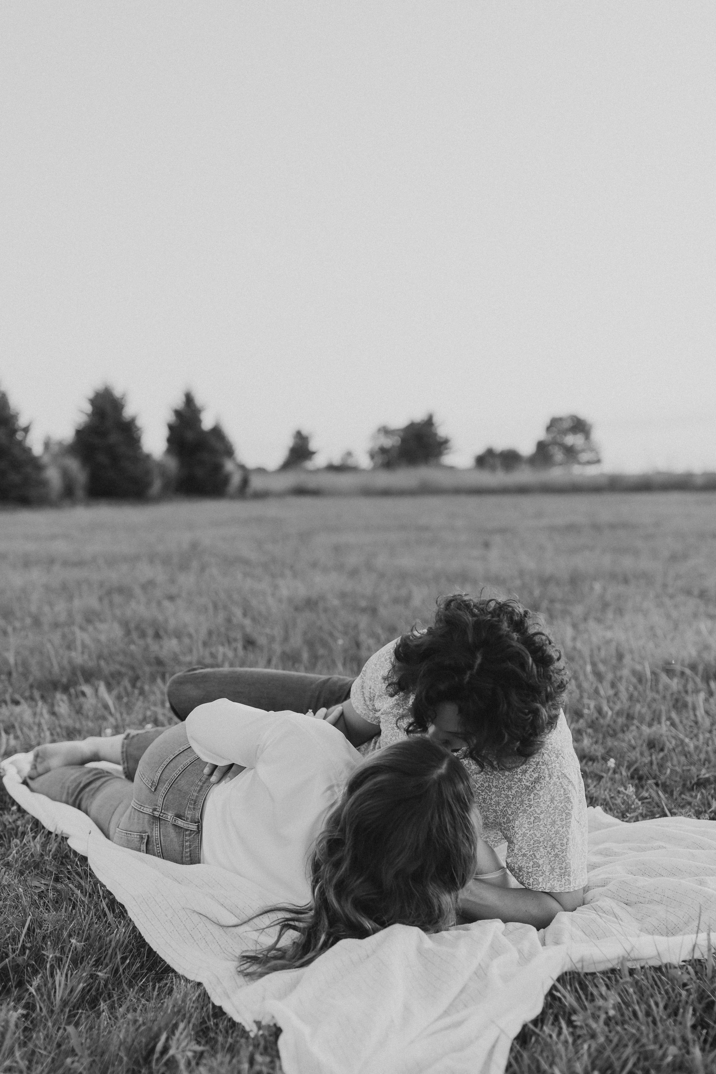  an engaged couple lay on a picnic blanket in a field as the sun sets 