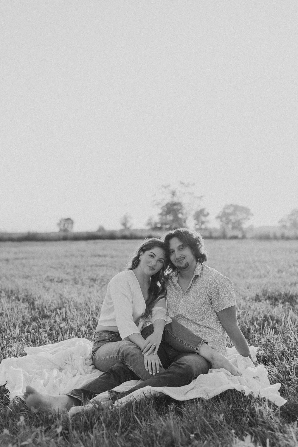  a newly engaged couple lean together as they sit on a picnic blanket in a field at sunset during a session with their engagement photographer 