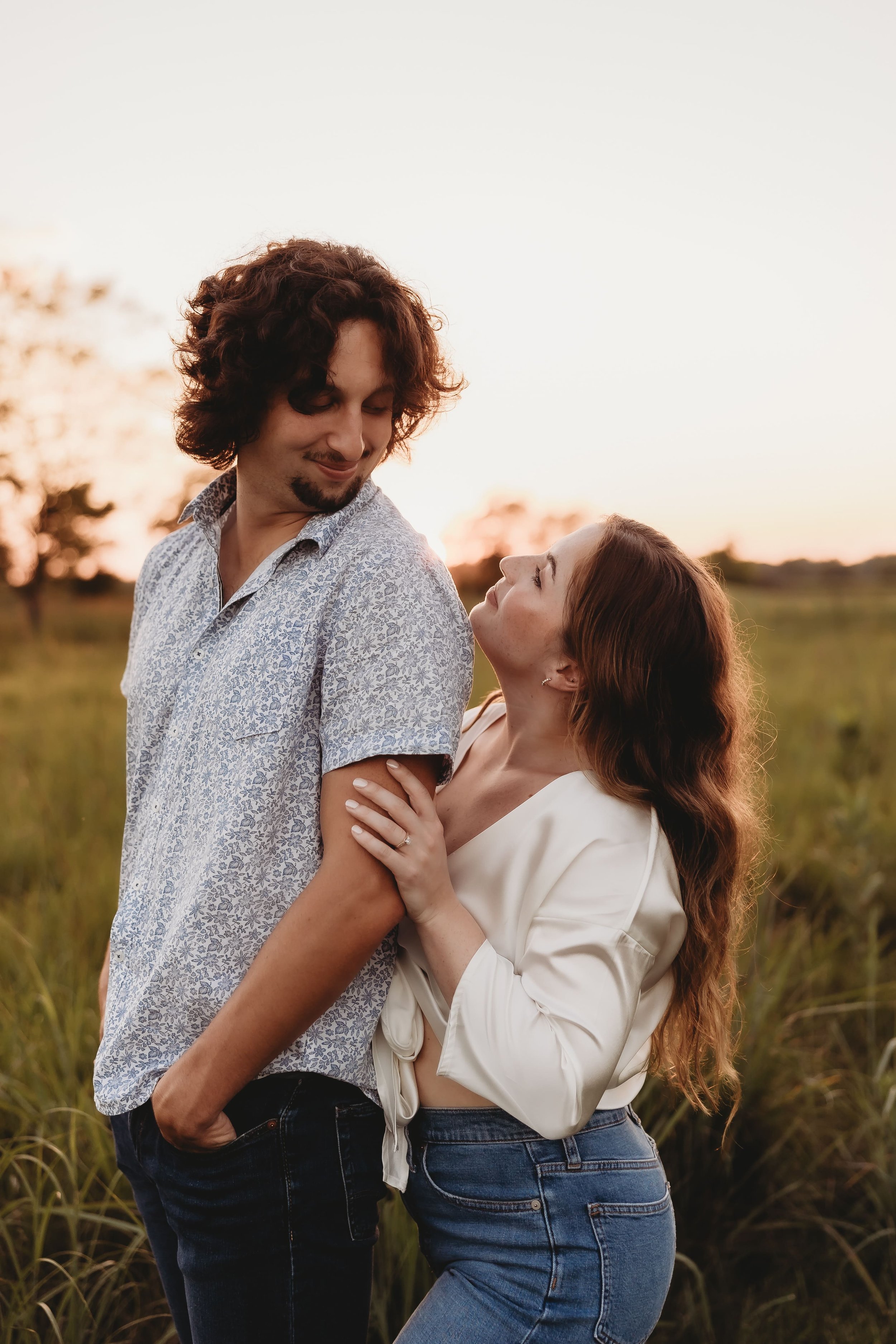  a couple smile at each other as they stand together in a field as the sun sets 