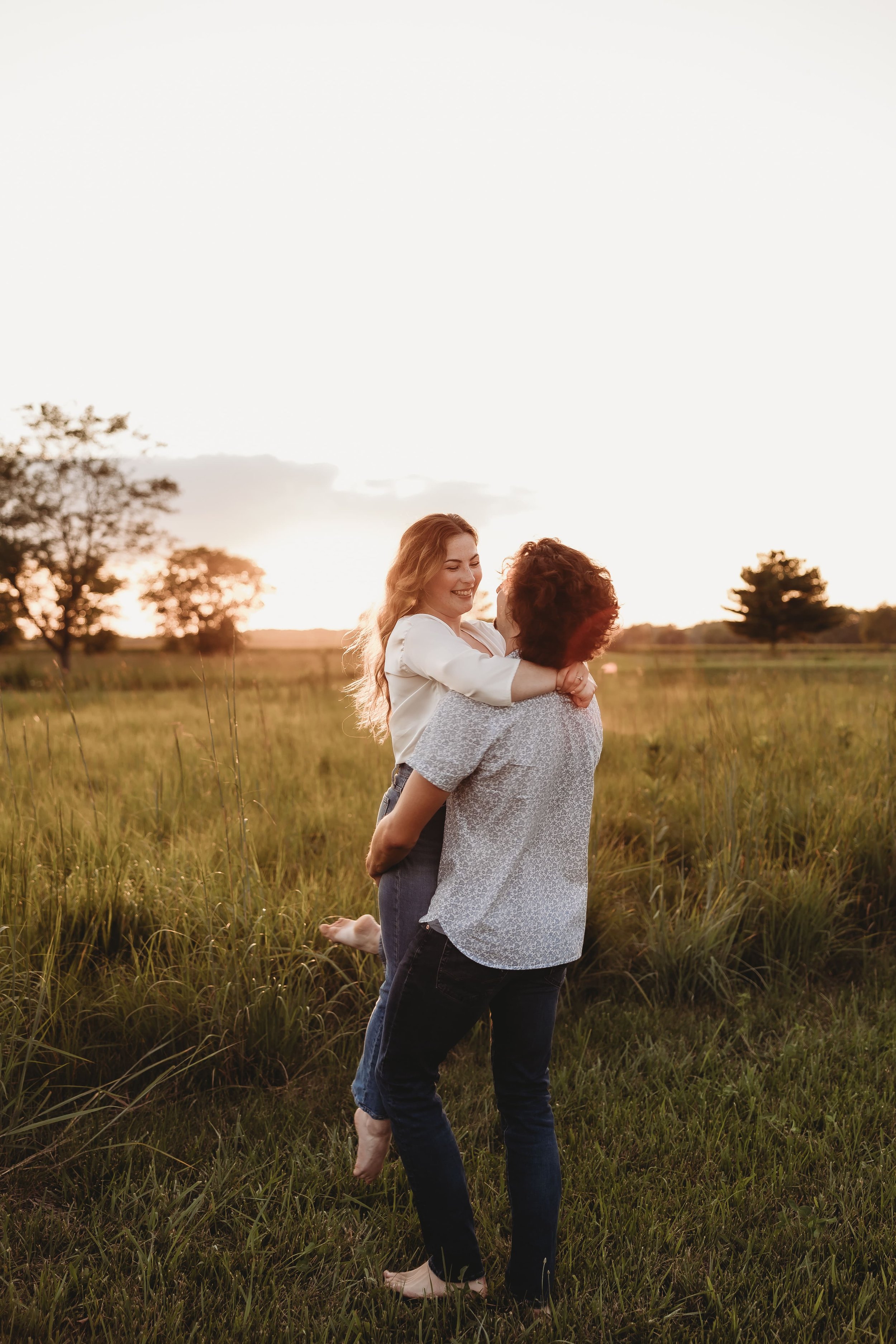 a man swings his fiancee around in a field as the sun sets behind them 