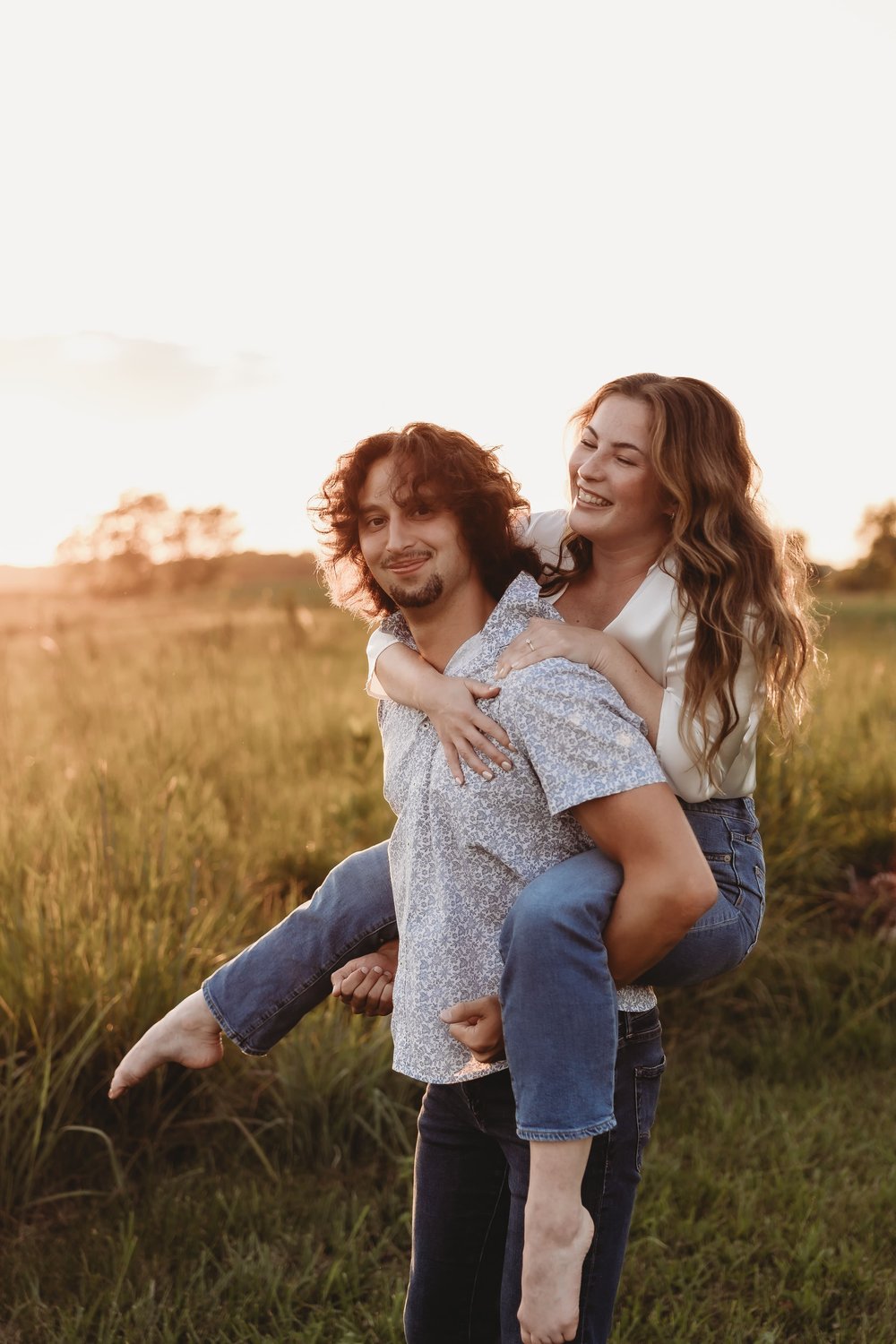  a man gives his fiancee a piggy back ride in a field as the sun sets 
