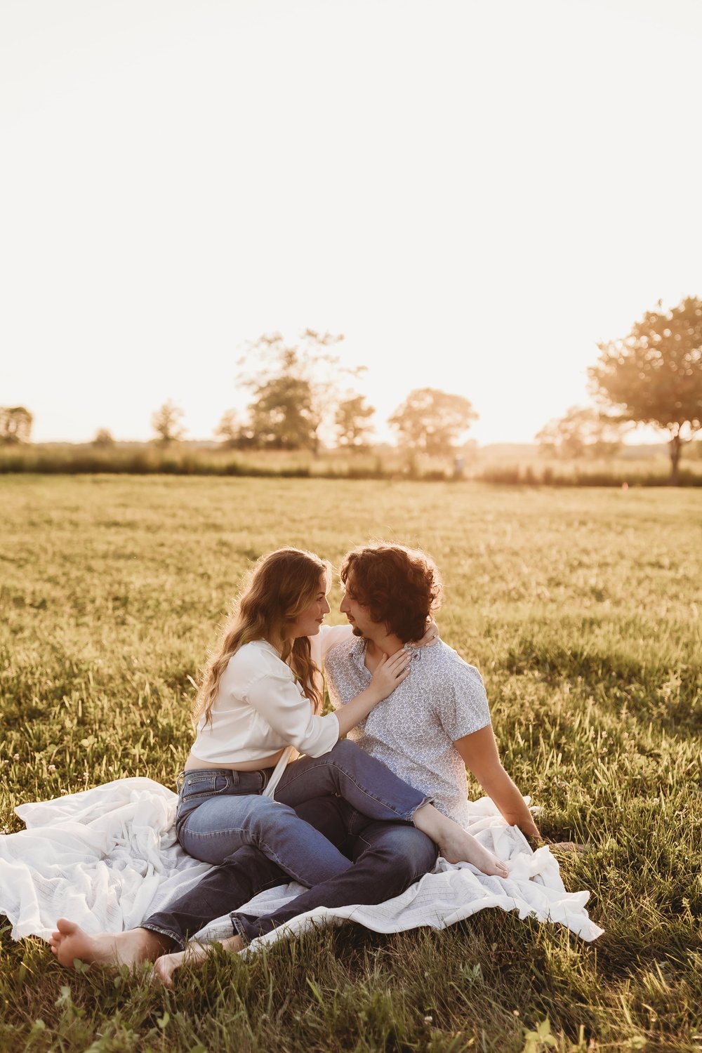  an engaged couple share a moment on a picnic blanket in a field as the sun sets behind them during their engagement photos 
