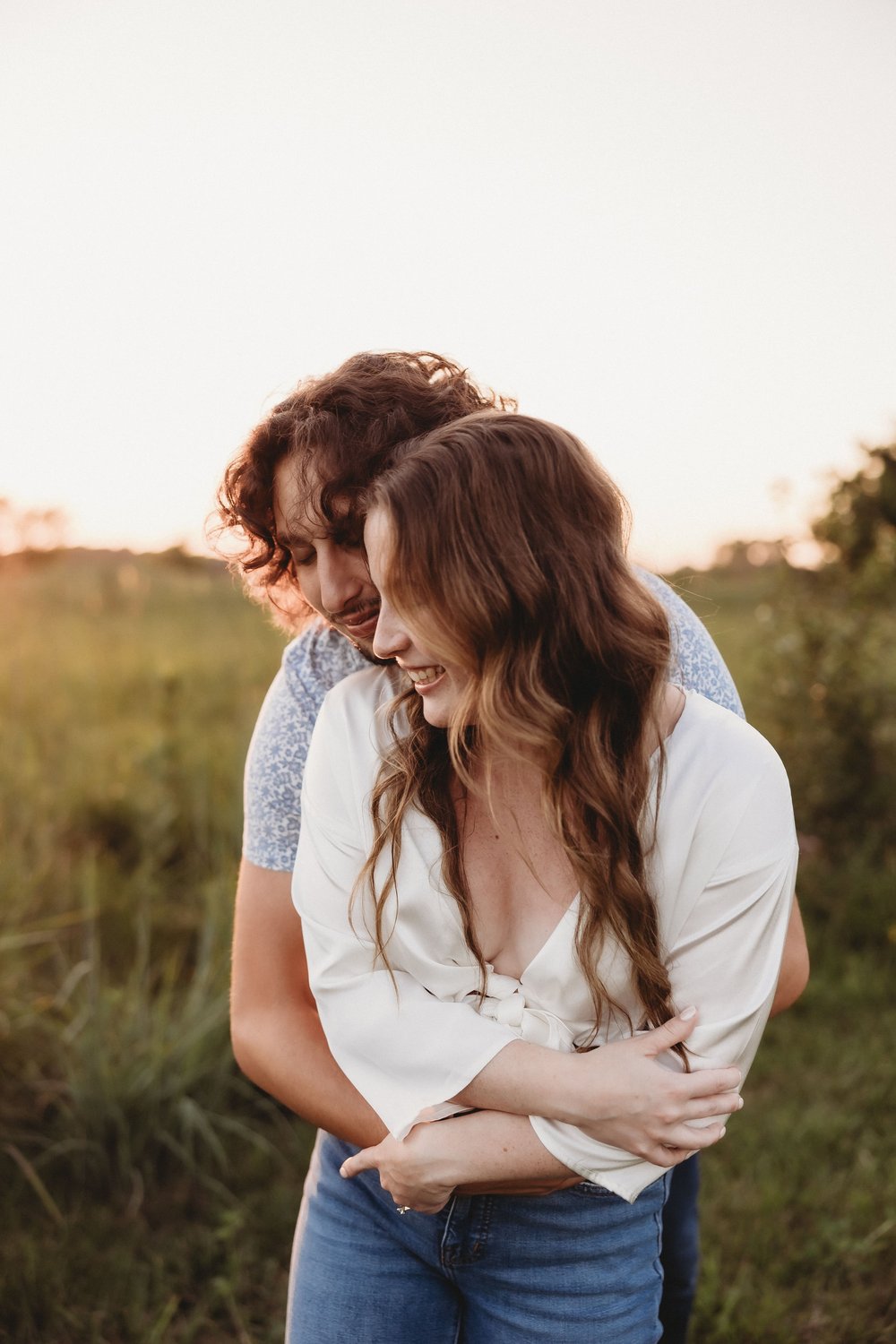  an engaged couple smile together in a field as the sun sets behind them for their engagement photos 