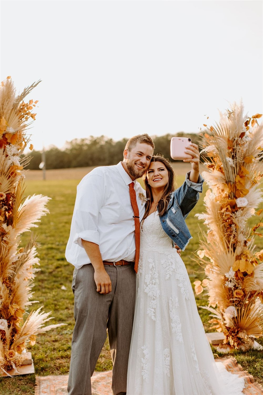  hannah and lance smile for a polaroid selfie in front of their boho dried floral wedding arbor 
