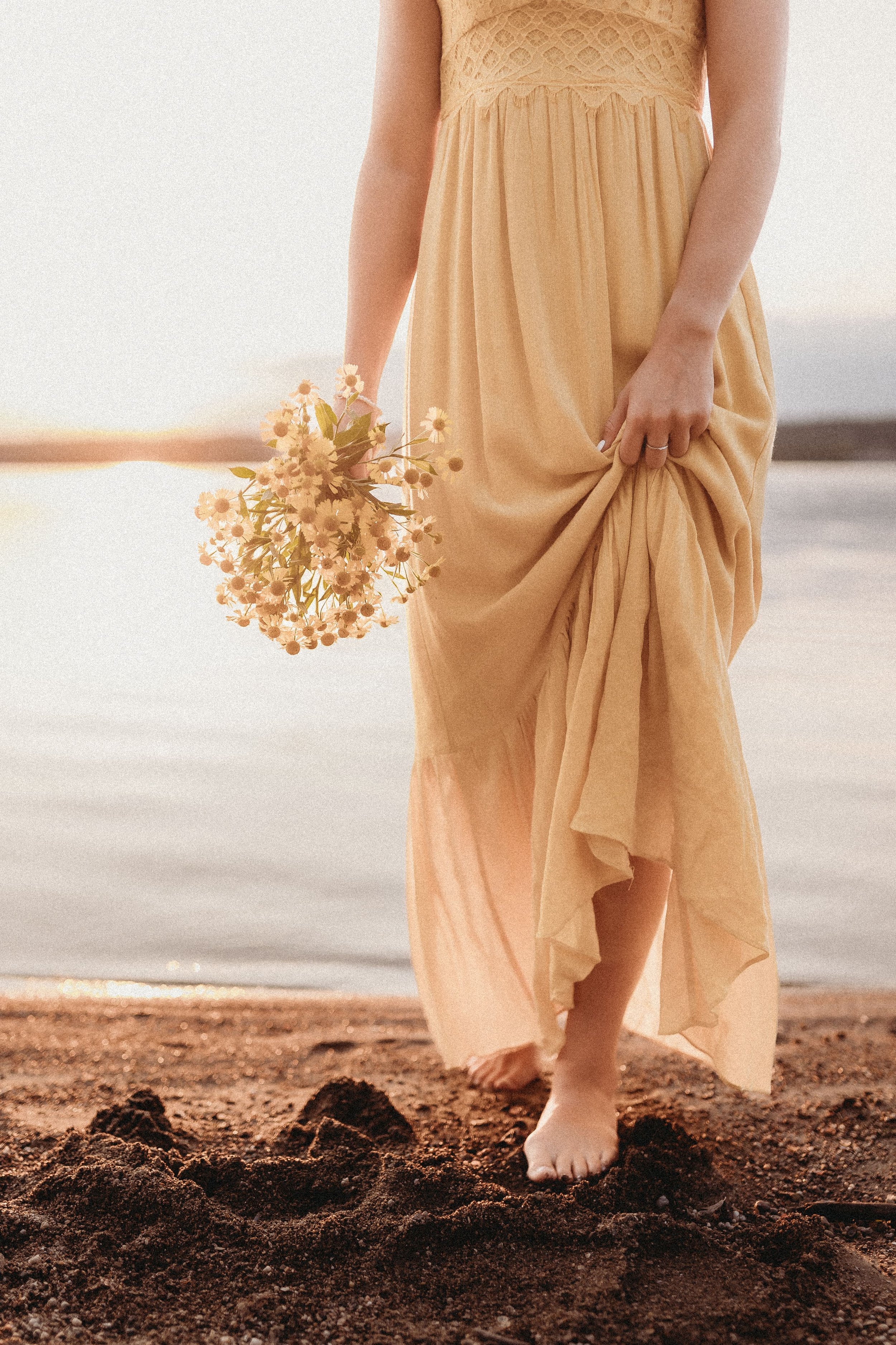  a woman wearing a long yellow dress stands barefoot on the banks of the river while holding a bunch of yellow flowers 