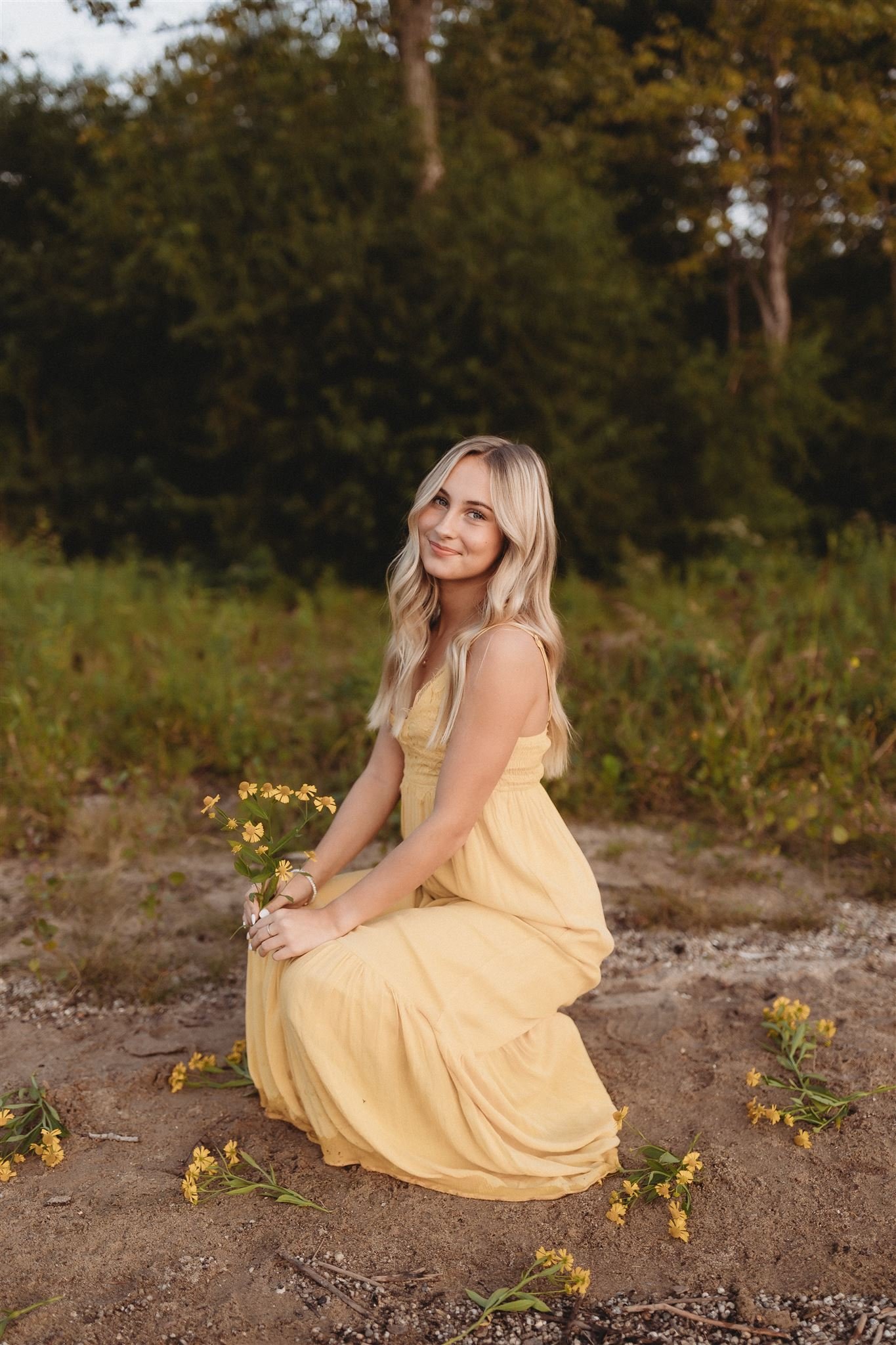  a young woman crouches down in a sandy/grassy area while wearing a long yellow dress 