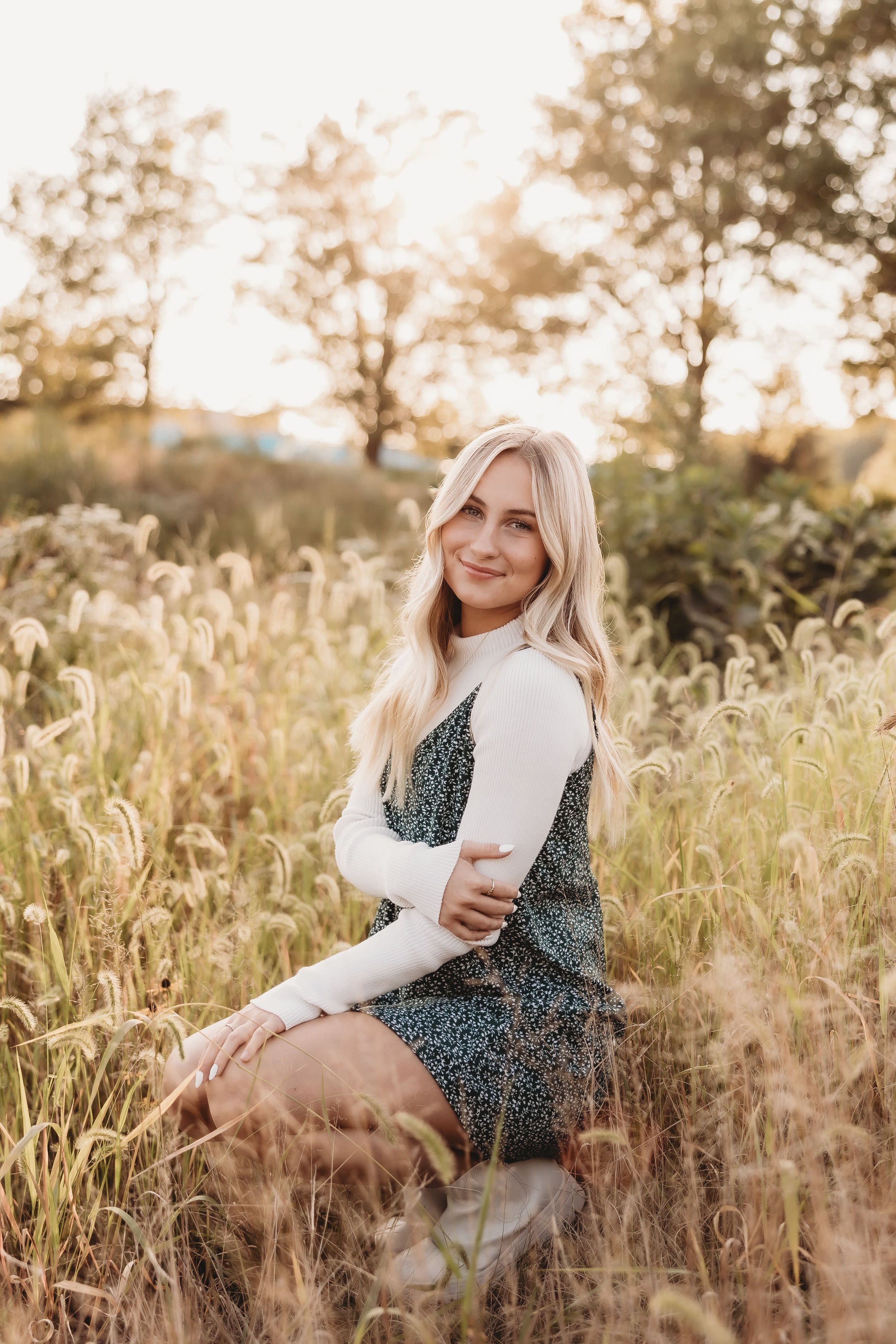  a young woman smiles as she crouches in a field as the sun sets behind her for her senior photos 