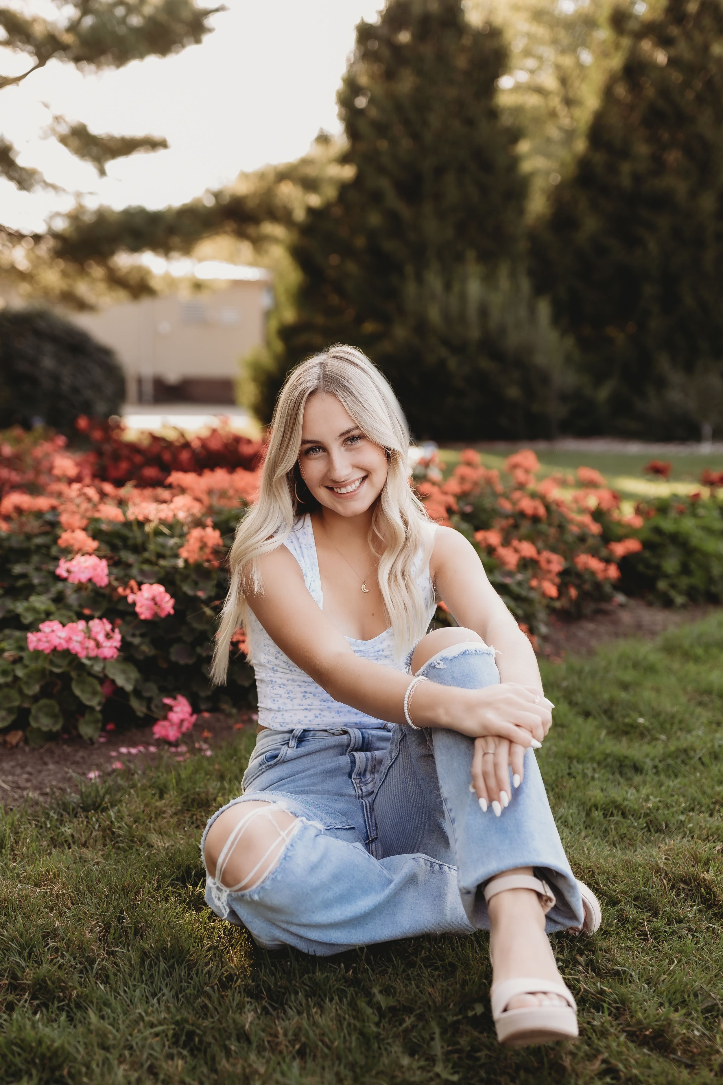  parker sits in the grass and smiles with her arms resting on her leg in front of a flower bed for her senior photos 