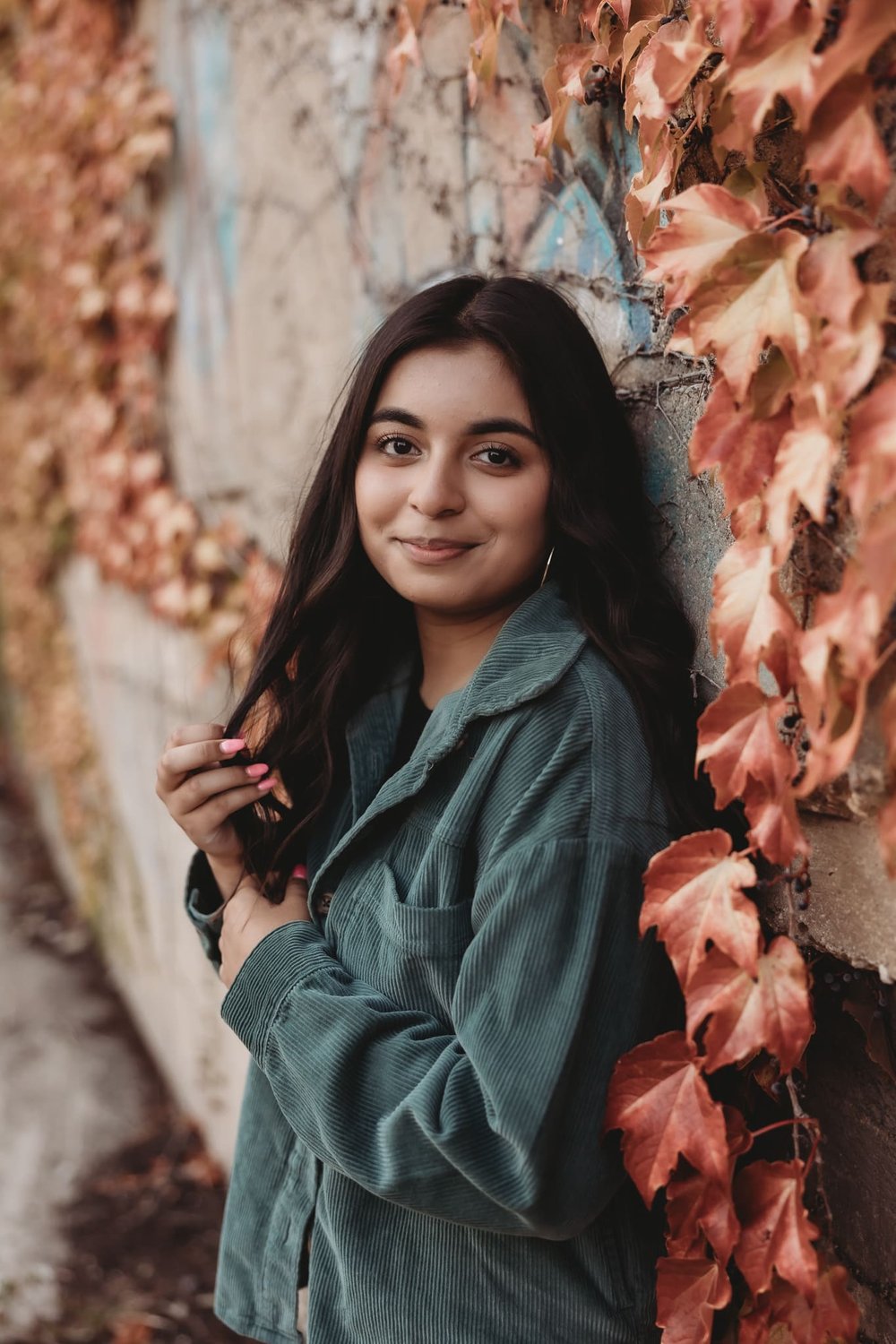  a young woman smiles leaning against a wall covered in climbing fall leaves for senior photography 