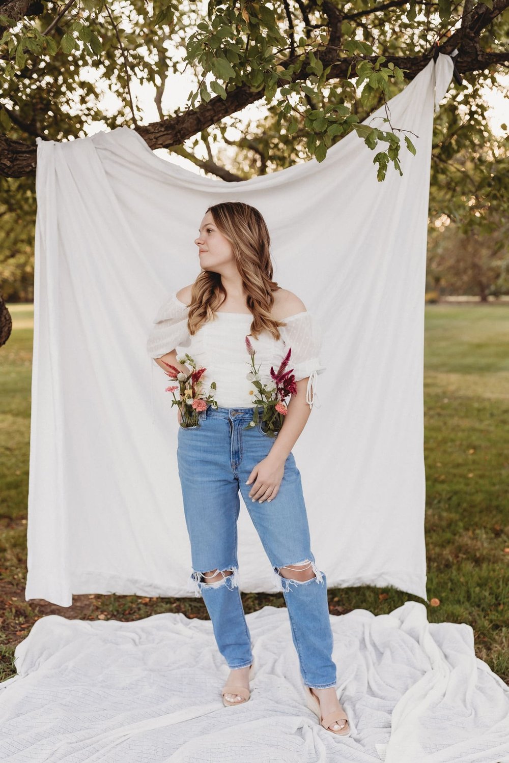  a girl stands in front of a sheet hanging from a tree with flowers in her pockets 