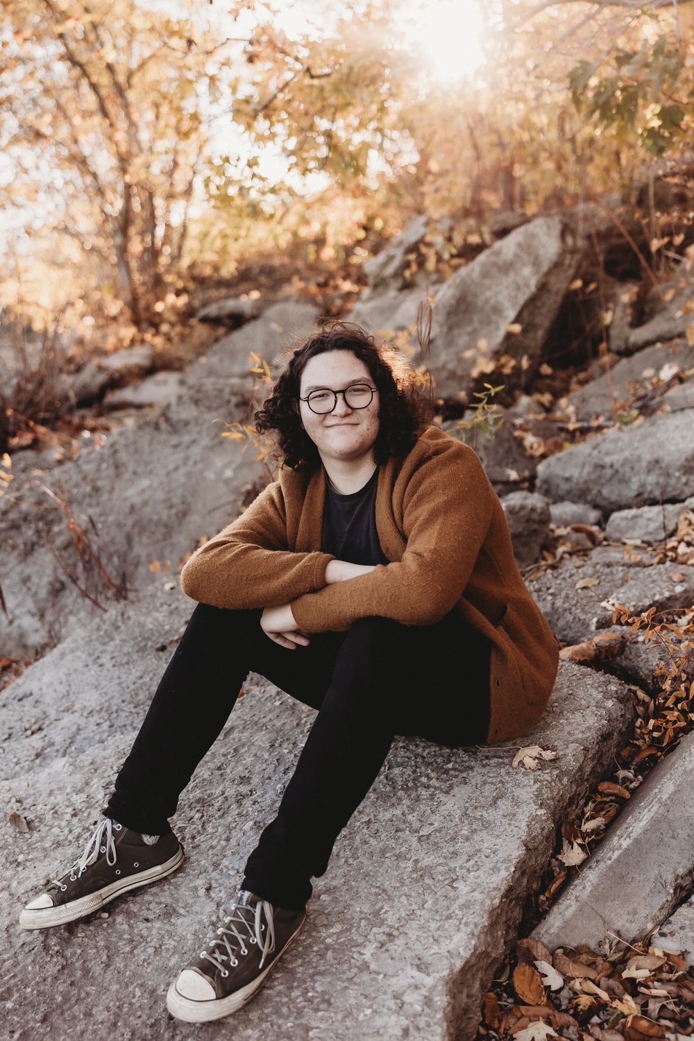  senior photography, a young man sits on a pile of rocks while wearing green converse, a dark outfit and a rust orange cardigan 