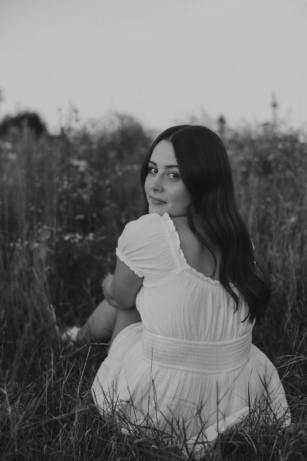  a girl wearing a white dress in a field looks over her shoulder and smiles for her senior photography 