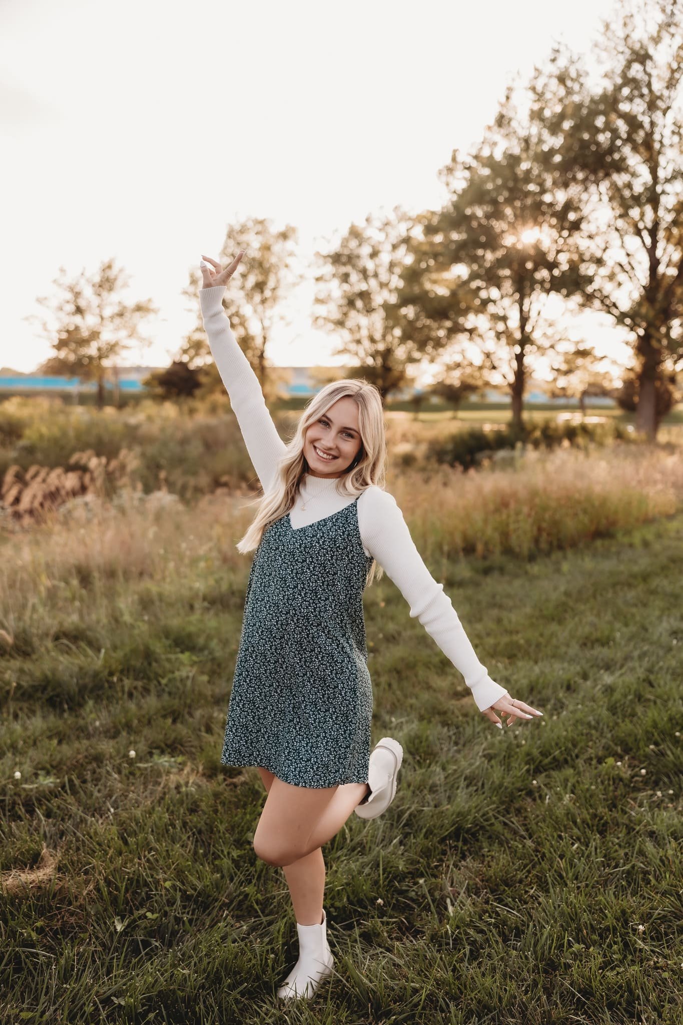  for senior photography, a young woman stands on one leg with her arms in the air in an open field  