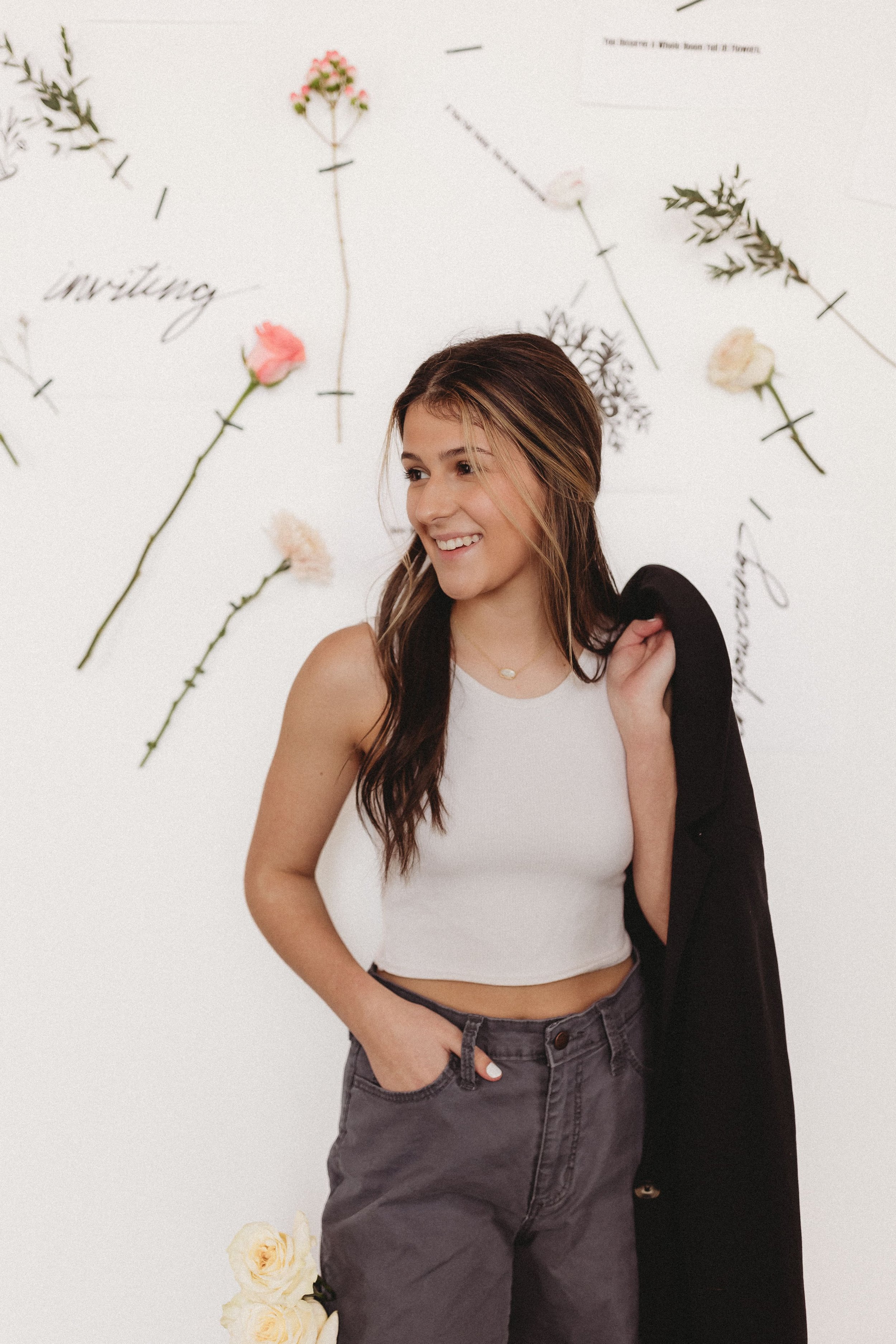  a girl smiles for business branding photography with a black jacket draped over her shoulder with flowers and branded prints taped to the white wall behind her 