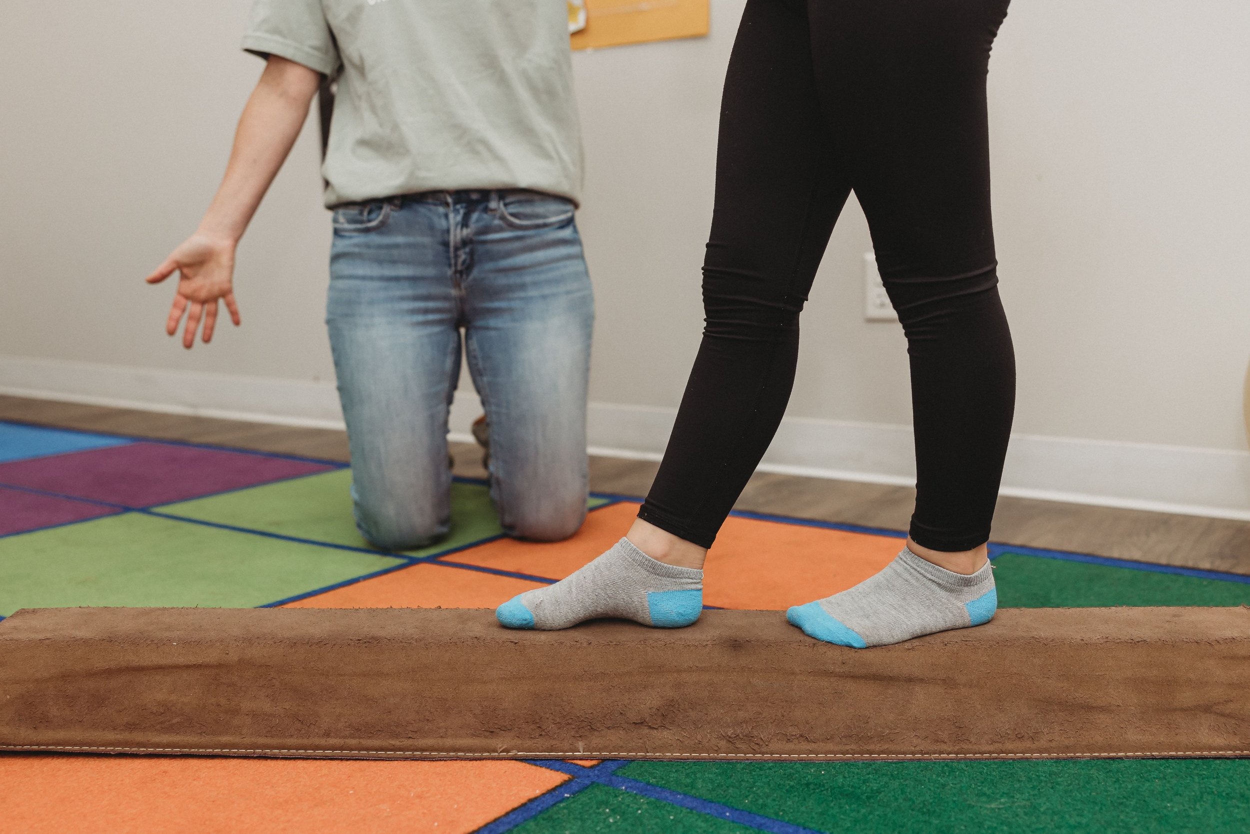  an adult kneels next to a balance beam that a child walks on in front of them 