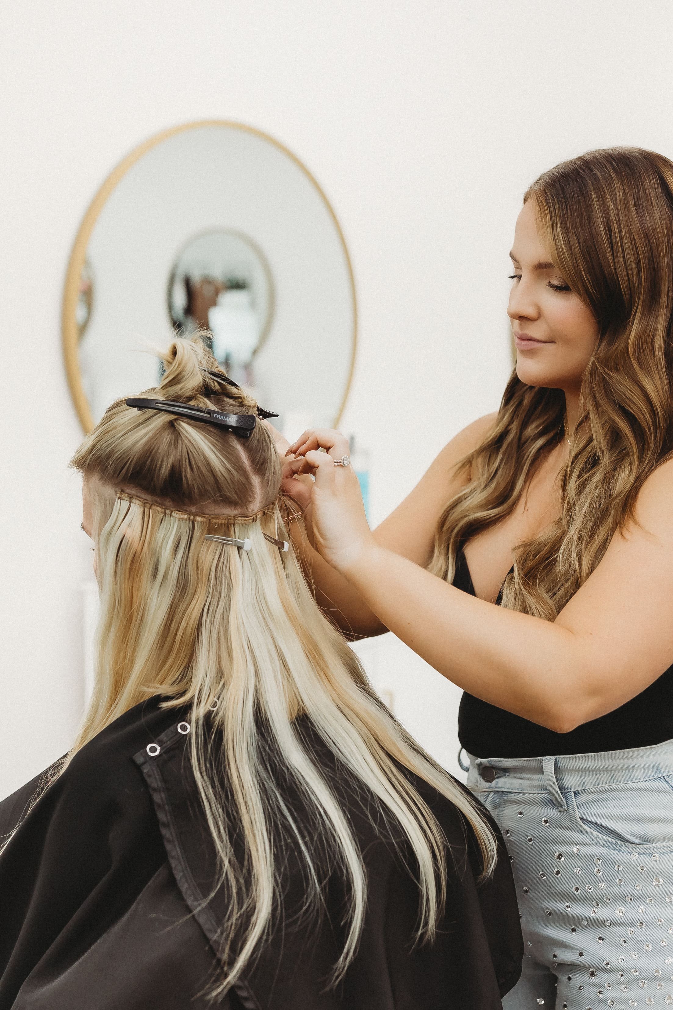  a hair stylist looks down at her client’s head as she puts in hair extensions 