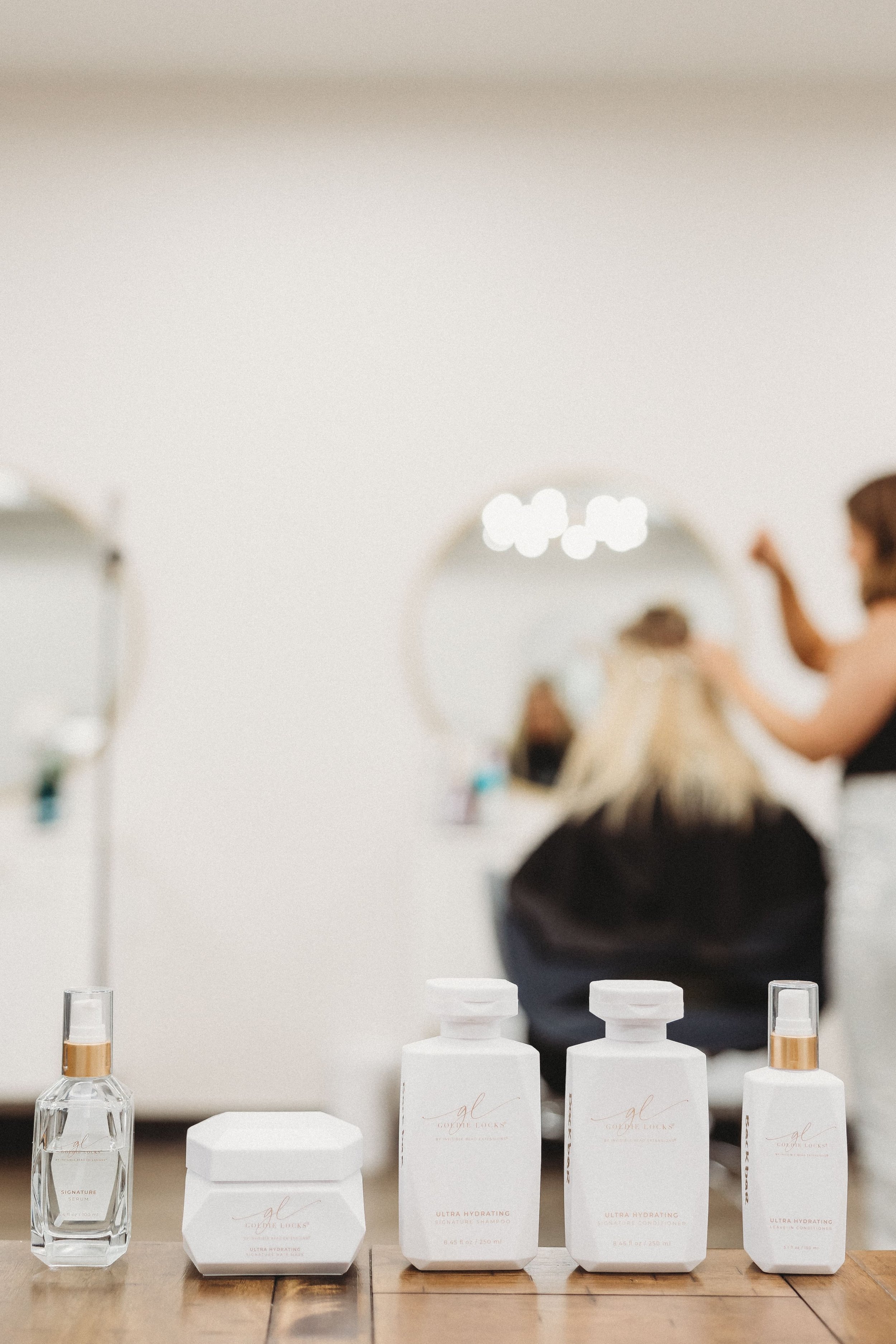  a collection of hair products stand in the foreground as a stylist puts extensions in her clients hair in the background 