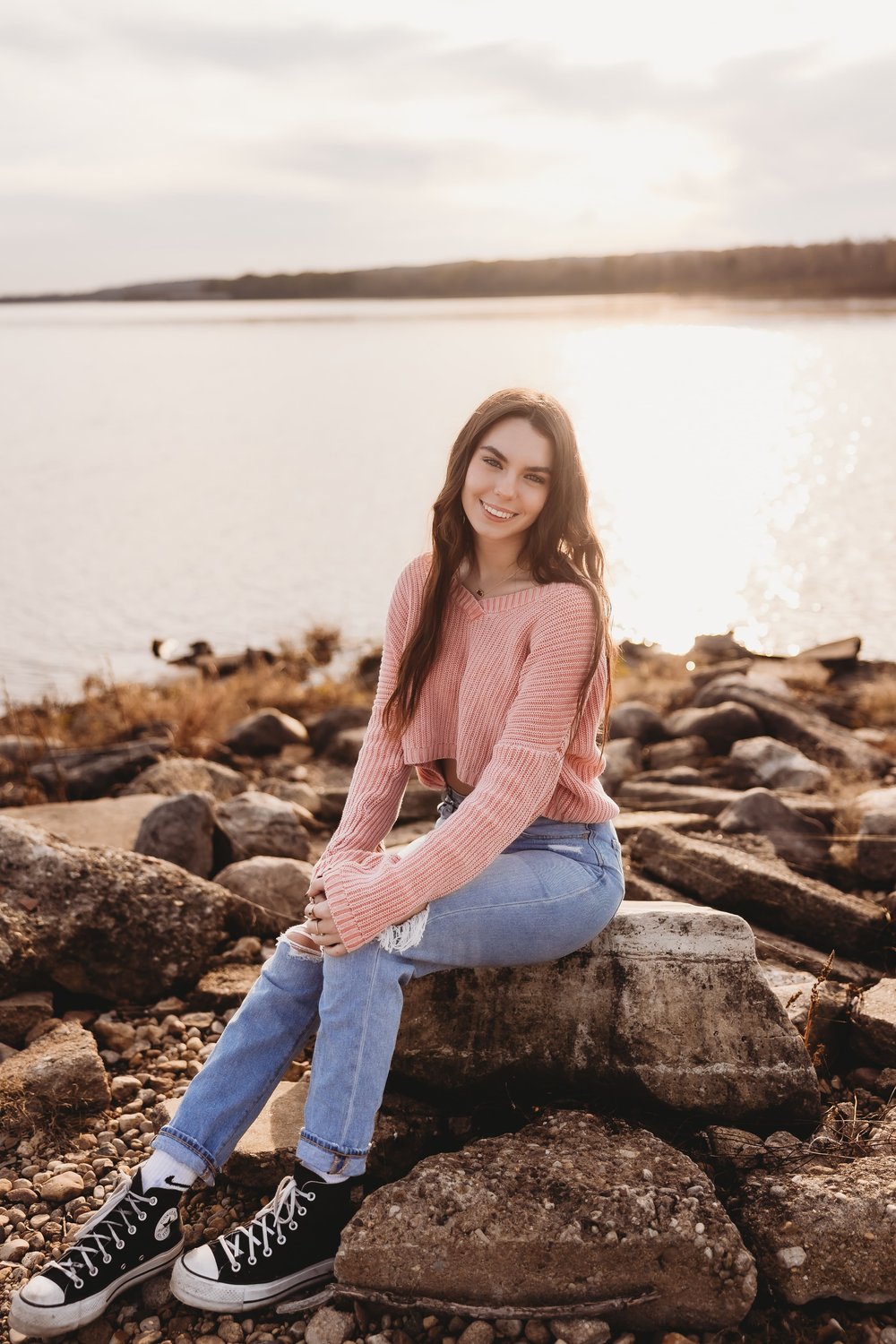  a senior girl sits on a rock in front of the illinois river and smiles with her head tilted to the side 