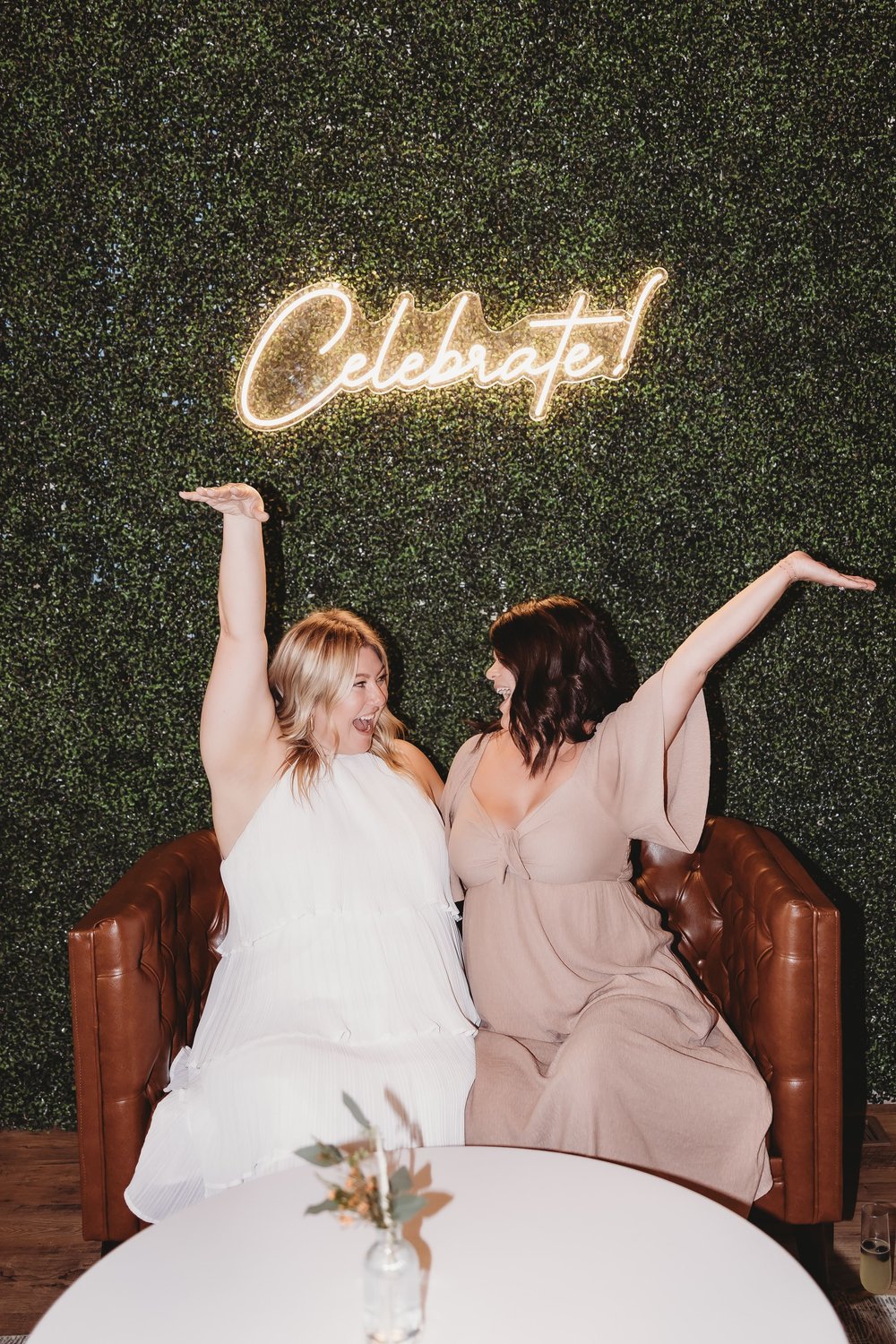  two women sit under the neon celebrate sign at parkside event room with their arms in the air and excited looks on their faces as they look at each other during brand photos for the event room   