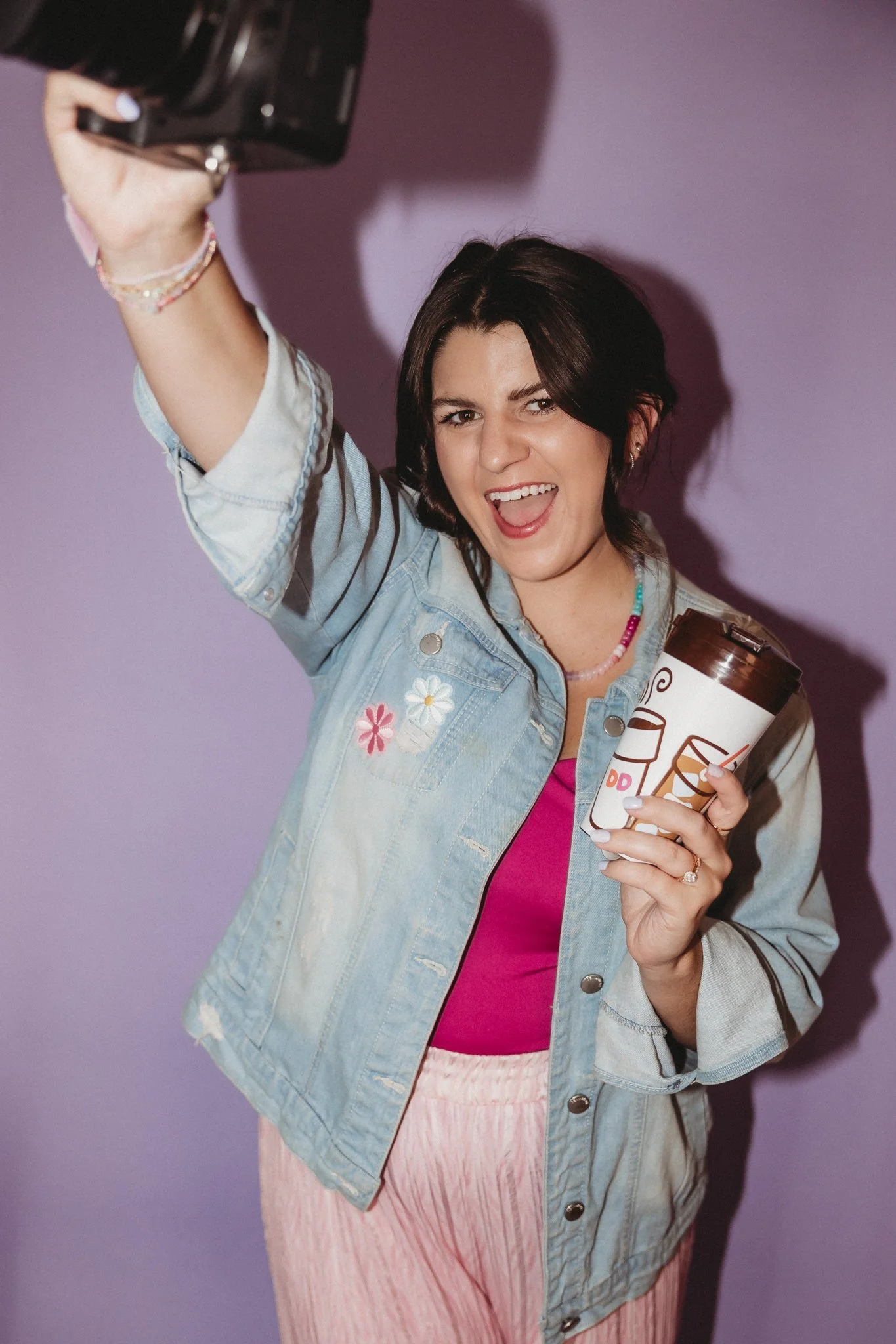 A young woman with short dark hair, smiling and winking, taking a selfie with a camera. She is wearing a light blue denim jacket with floral embroidery, a pink top, and pink pants, standing against a lavender background. She holds a disposable coffee cup in her left hand.