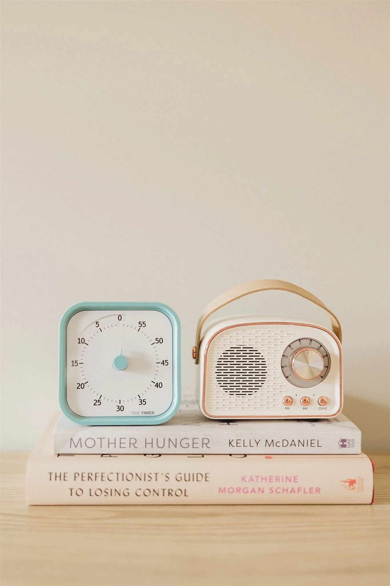 A light blue kitchen timer and a vintage radio on top of two books, "Mother Hunger" by Kelly McDaniel and "The Perfectionist's Guide to Losing Control" by Katherine Morgan Schafler, on a wooden surface against a beige wall.