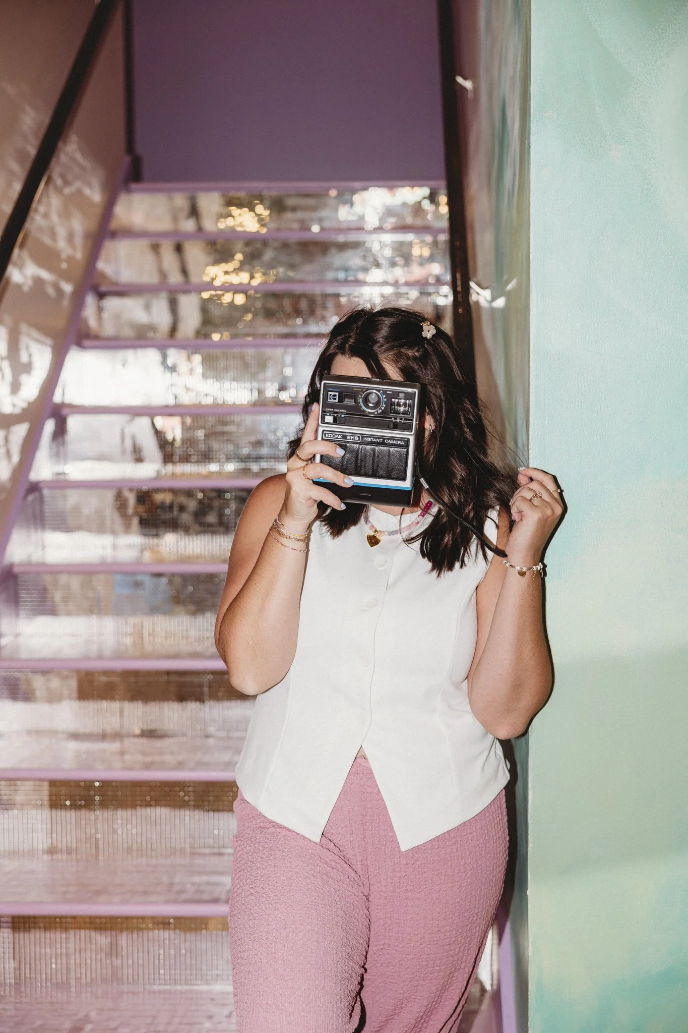 A woman with dark hair, wearing a white sleeveless top and pink textured pants, holding a vintage instant camera up to her face. She stands on a staircase with reflective steps and a pastel green wall to her right.