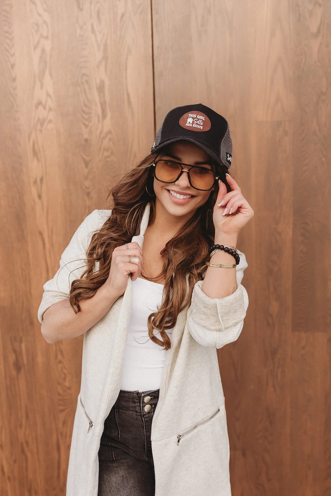 A smiling woman with long, wavy hair wearing large sunglasses, a black trucker hat, a white top, a light-colored jacket, and dark pants, standing in front of a wooden wall.