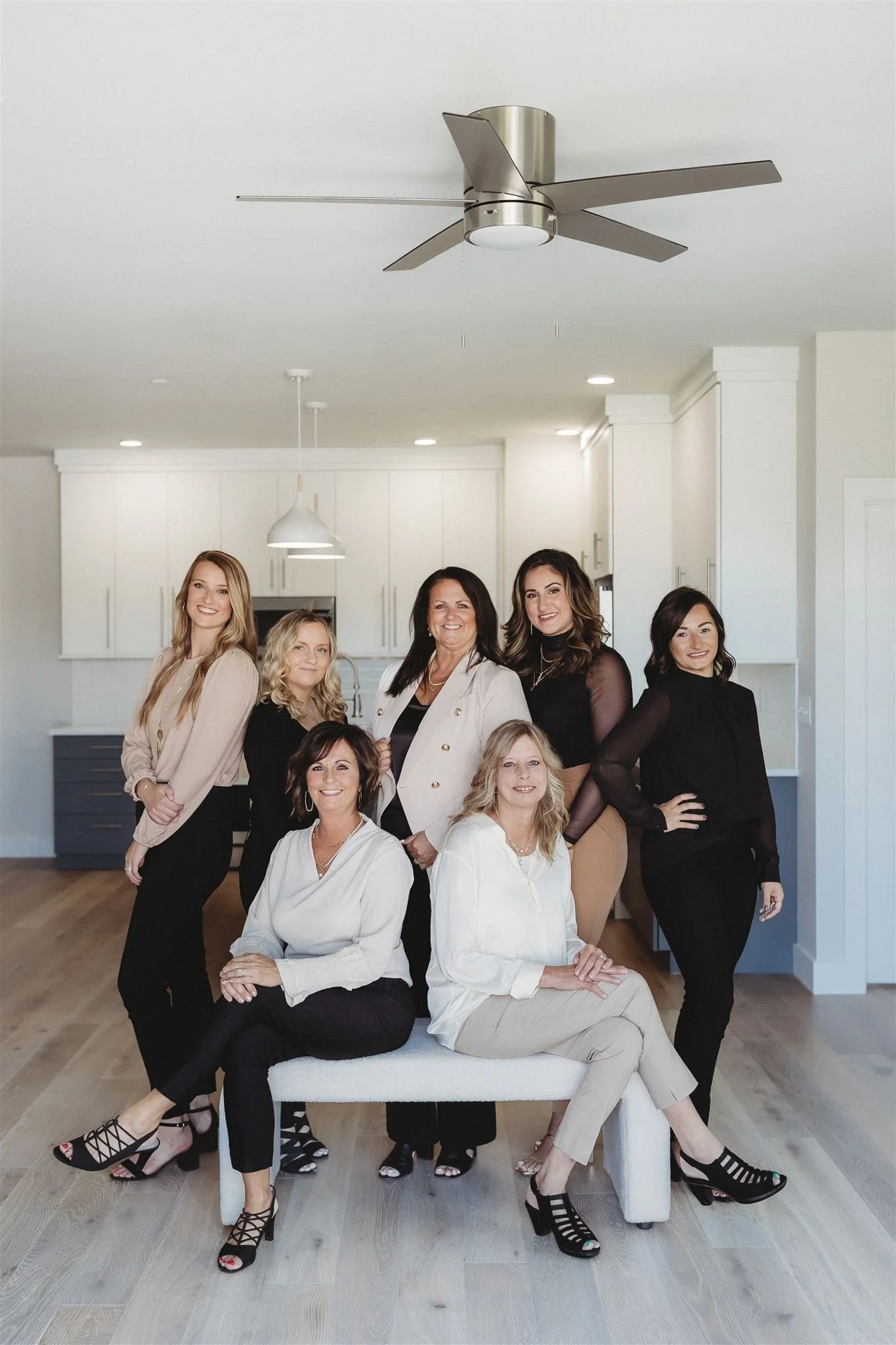 A group of eight women in a modern, white kitchen, posing for a photo, with some sitting and others standing, dressed in professional attire.