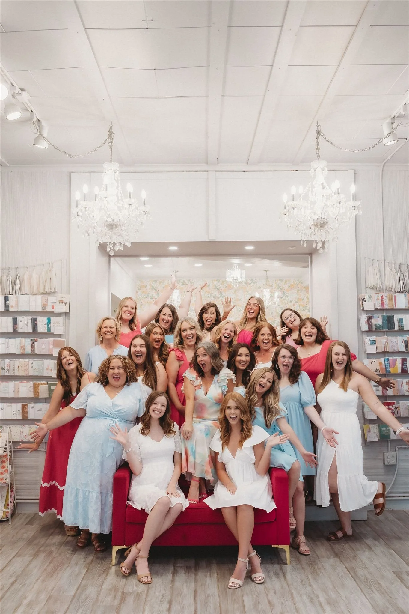 Group of women in colorful dresses celebrating in a bright, elegantly decorated room with chandeliers and a bookshelf.