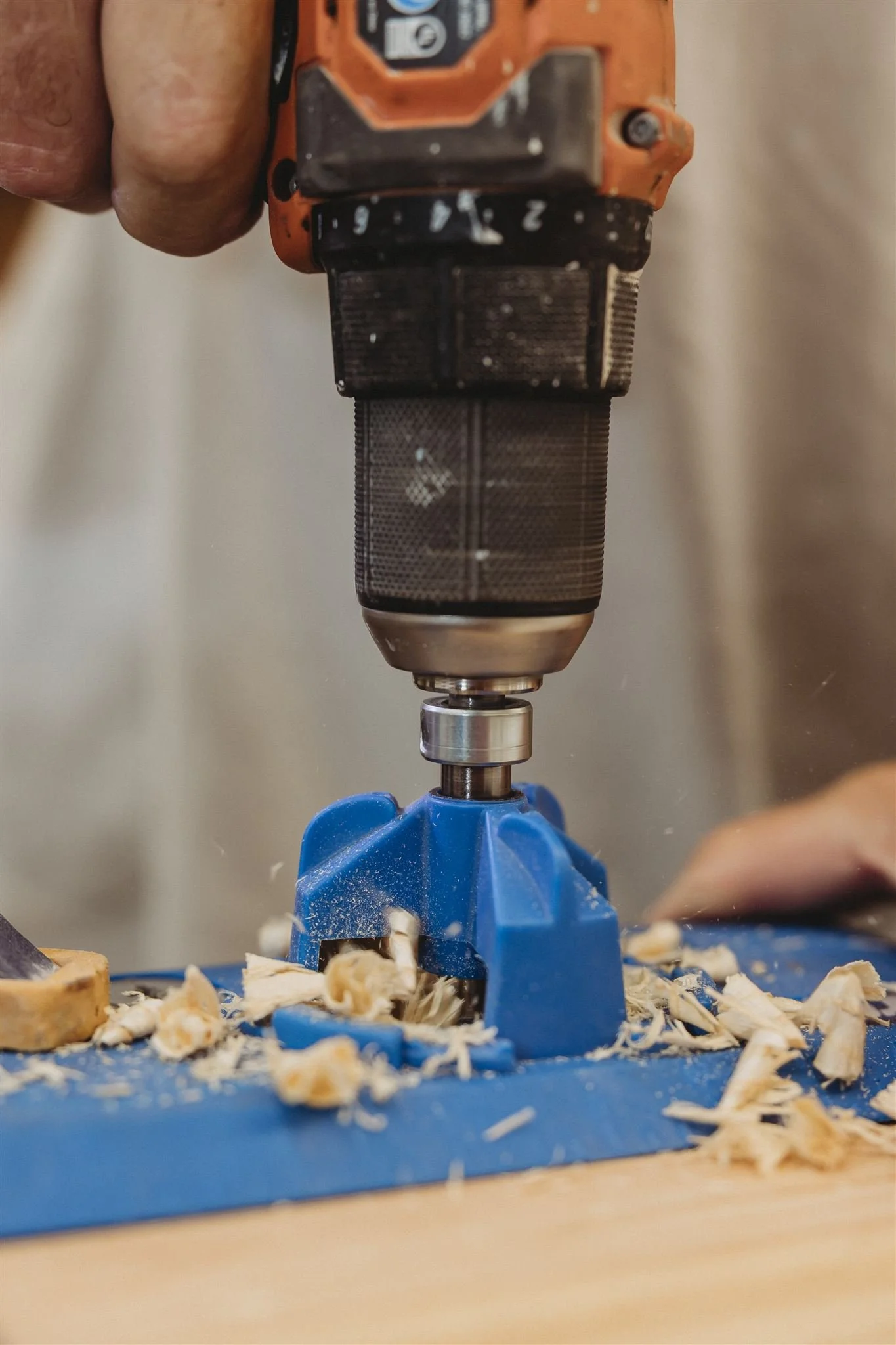 A close-up of a power drill drilling into a piece of wood, with wood shavings scattered around on a blue work surface.