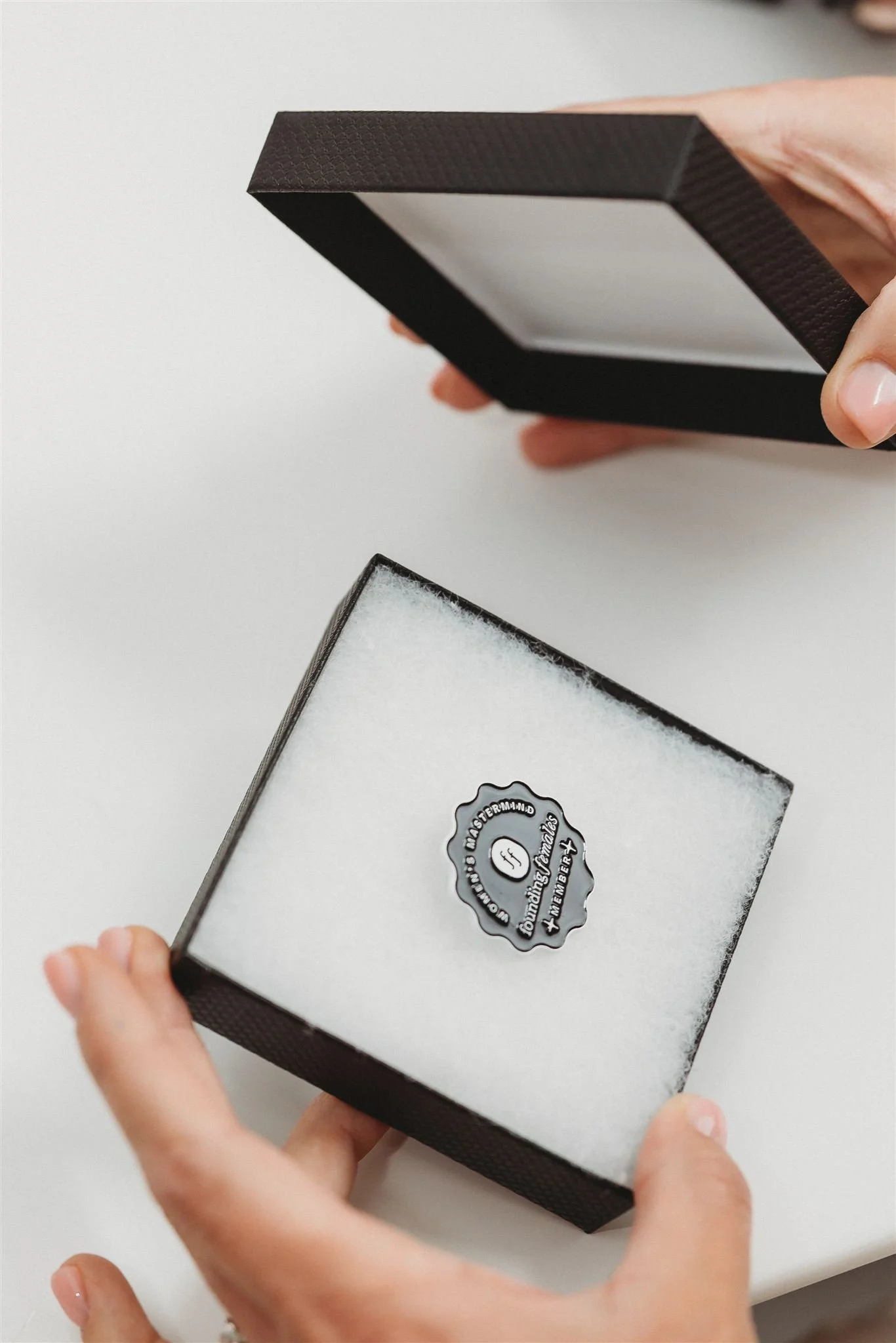 Hands holding an open black jewelry box with white interior, revealing a black sticker inside that reads 'The Founding Members.'