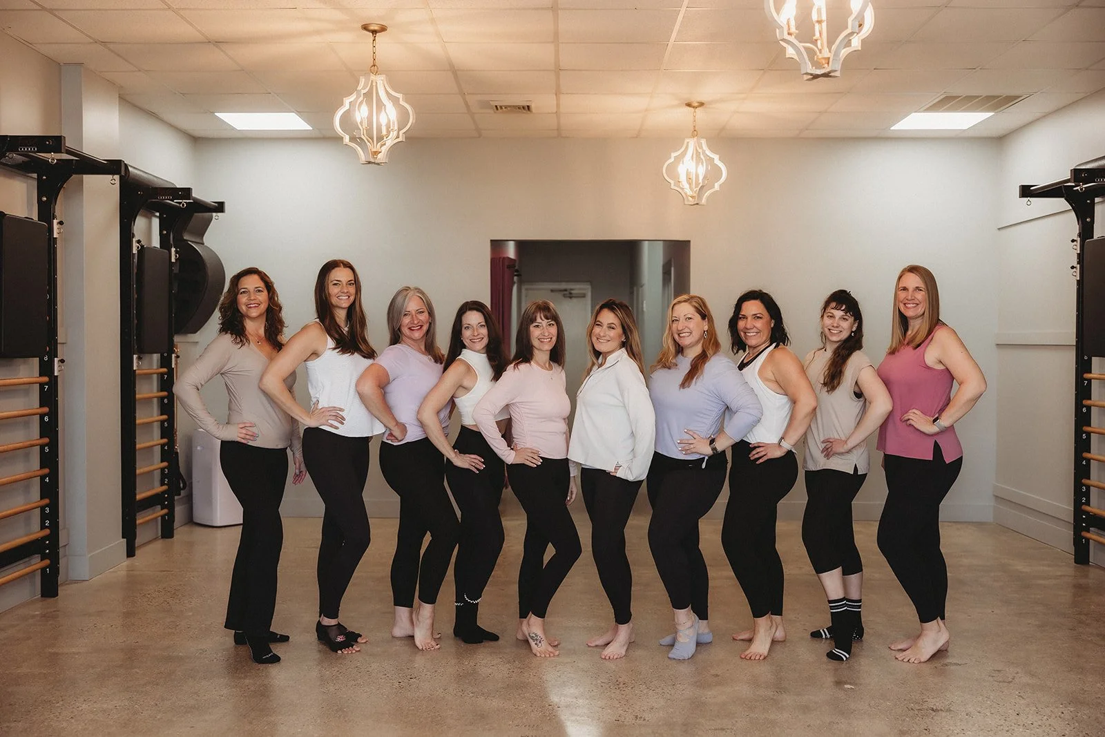 Group of eleven women standing in a line in a dance or fitness studio with wooden wall bars and chandeliers, smiling.
