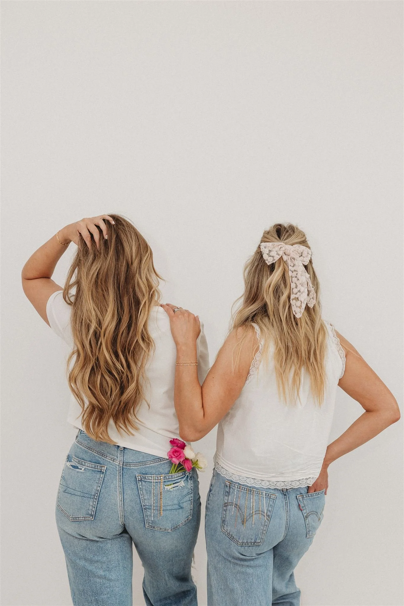 Two women with long blonde hair, dressed in white tops and jeans, facing away from the camera, standing against a plain white background.