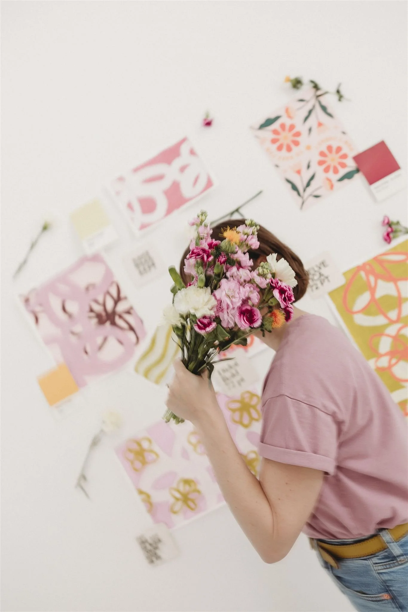 Person holding a bouquet of pink and white flowers, standing in front of a wall decorated with various colorful floral and abstract art prints.