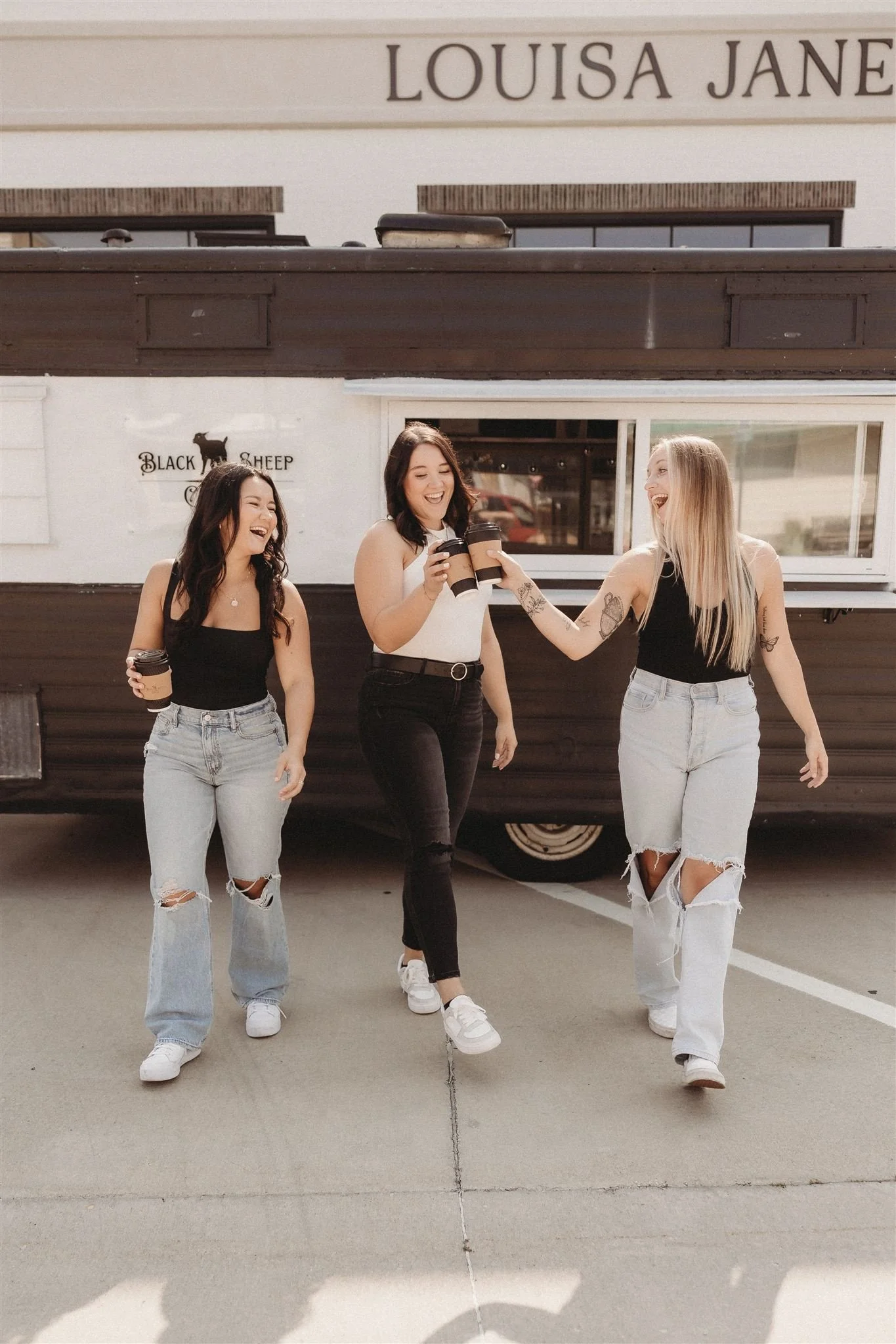 Three women standing together outside a food truck labeled Louisa Jane, holding coffee cups and laughing.