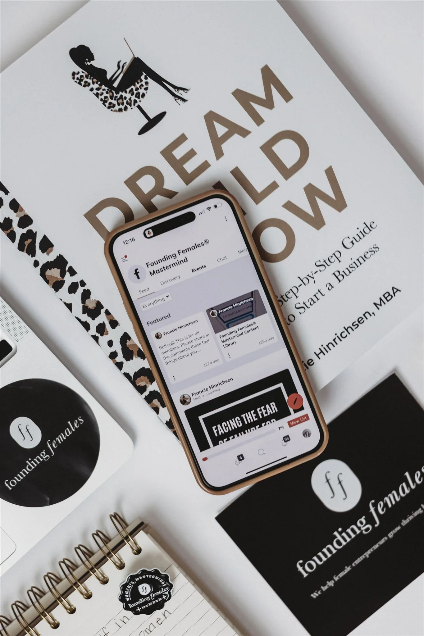 A smartphone displaying a Facebook group called 'Founding Females® Mastermind' is on a white desk surrounded by printed materials including a guidebook, stickers, and a notebook, all related to founding and growing a business.
