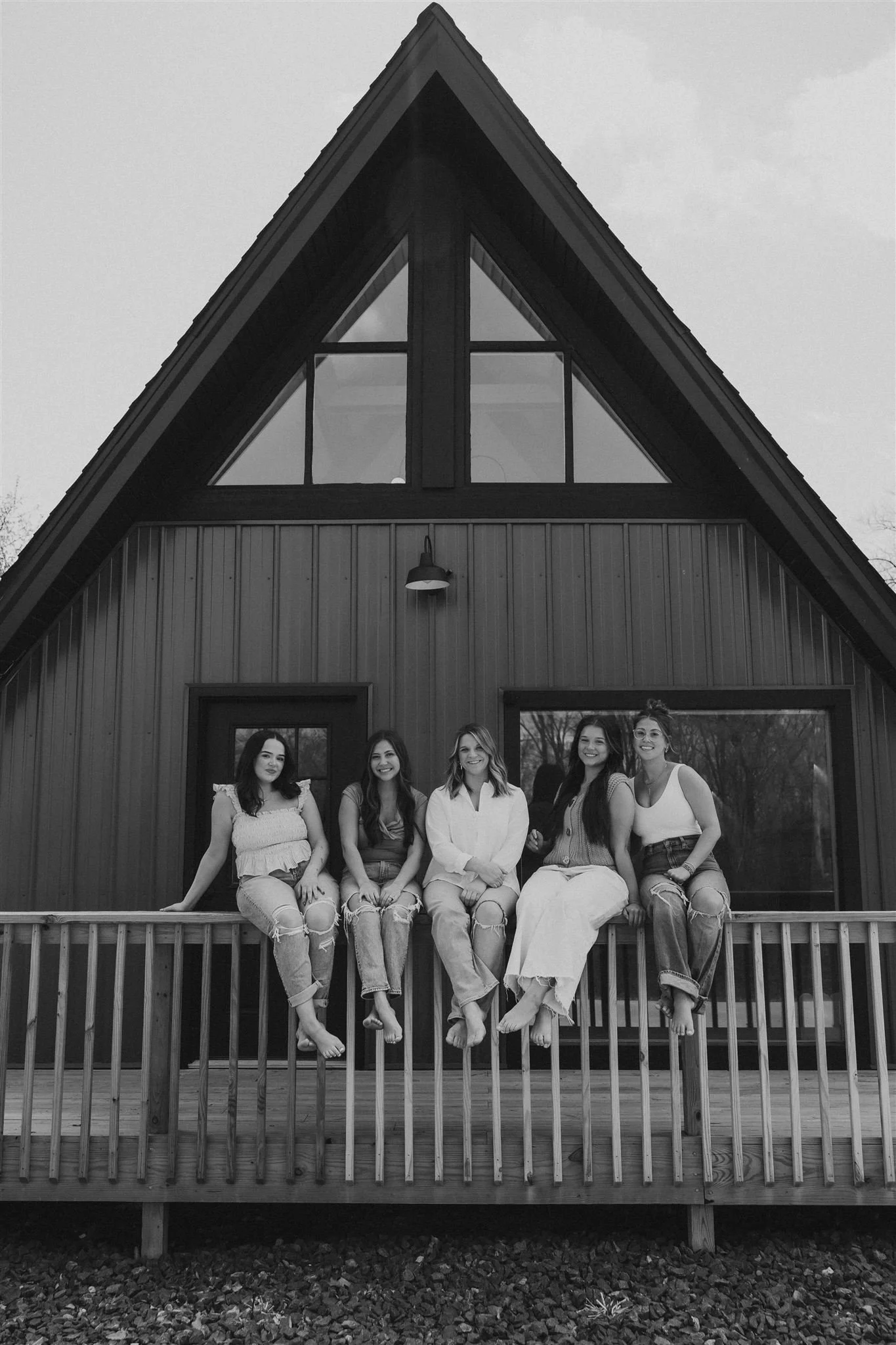 Five women sitting on a wooden balcony outside a modern house with a triangular roof and large windows, in black and white.