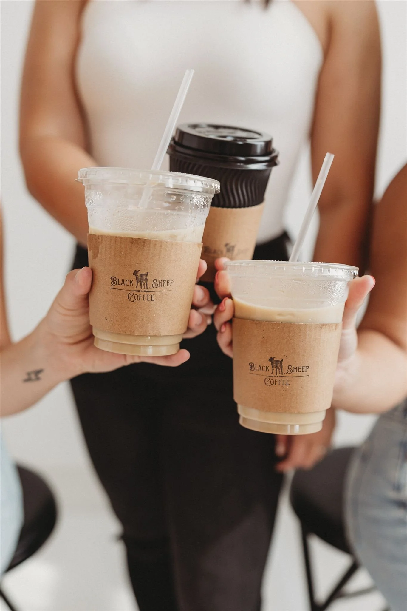 Three people holding glasses of coffee from Black Sheep Coffee, two with iced coffee and one with hot coffee, in a friendly and casual setting.