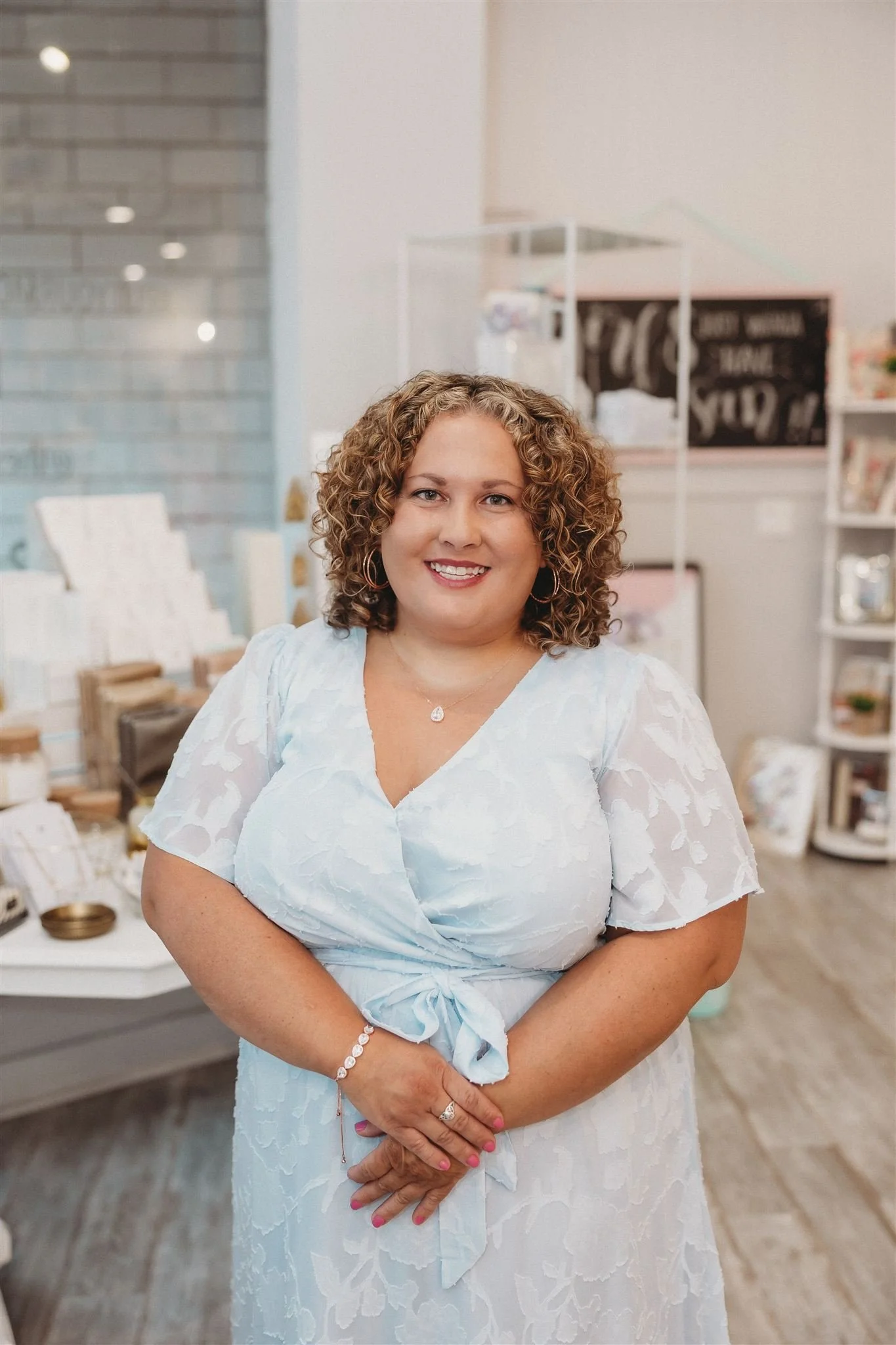 A woman with curly brown hair smiling in a store with white and pastel-colored decor.