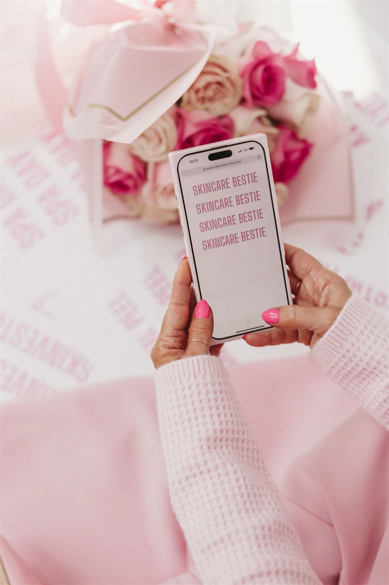 A person with pink nail polish and a pink sweater holding a smartphone with the screen displaying the words 'SKINCARE BESTIE' repeated four times. In the background, there is a pink gift box with a bow and a bouquet of pink and white roses, all on pink-themed wrapping paper.