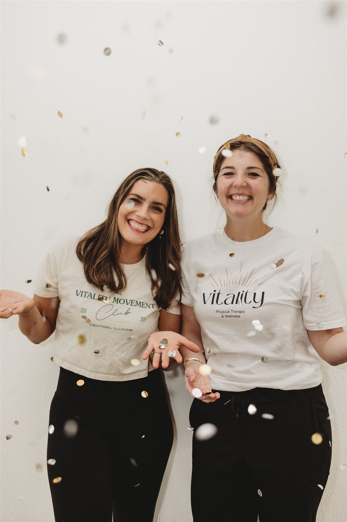Two smiling Vitality Physical Therapy and Wellness team members celebrating with confetti, wearing branded Vitality Movement Club and Vitality Physical Therapy t-shirts