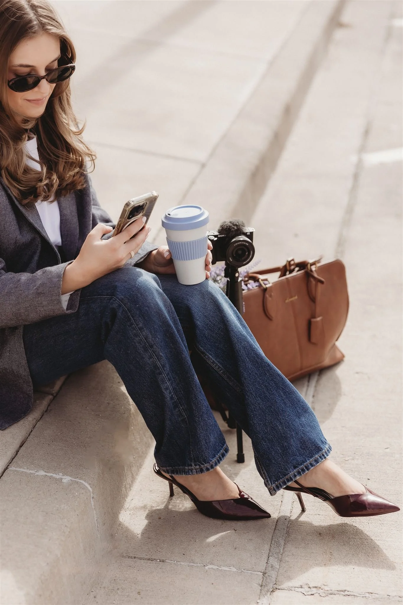 A woman sitting on a sidewalk, wearing sunglasses and heels, looking at her phone, with a coffee cup in hand, surrounded by a camera, a brown leather bag, and a tripod.