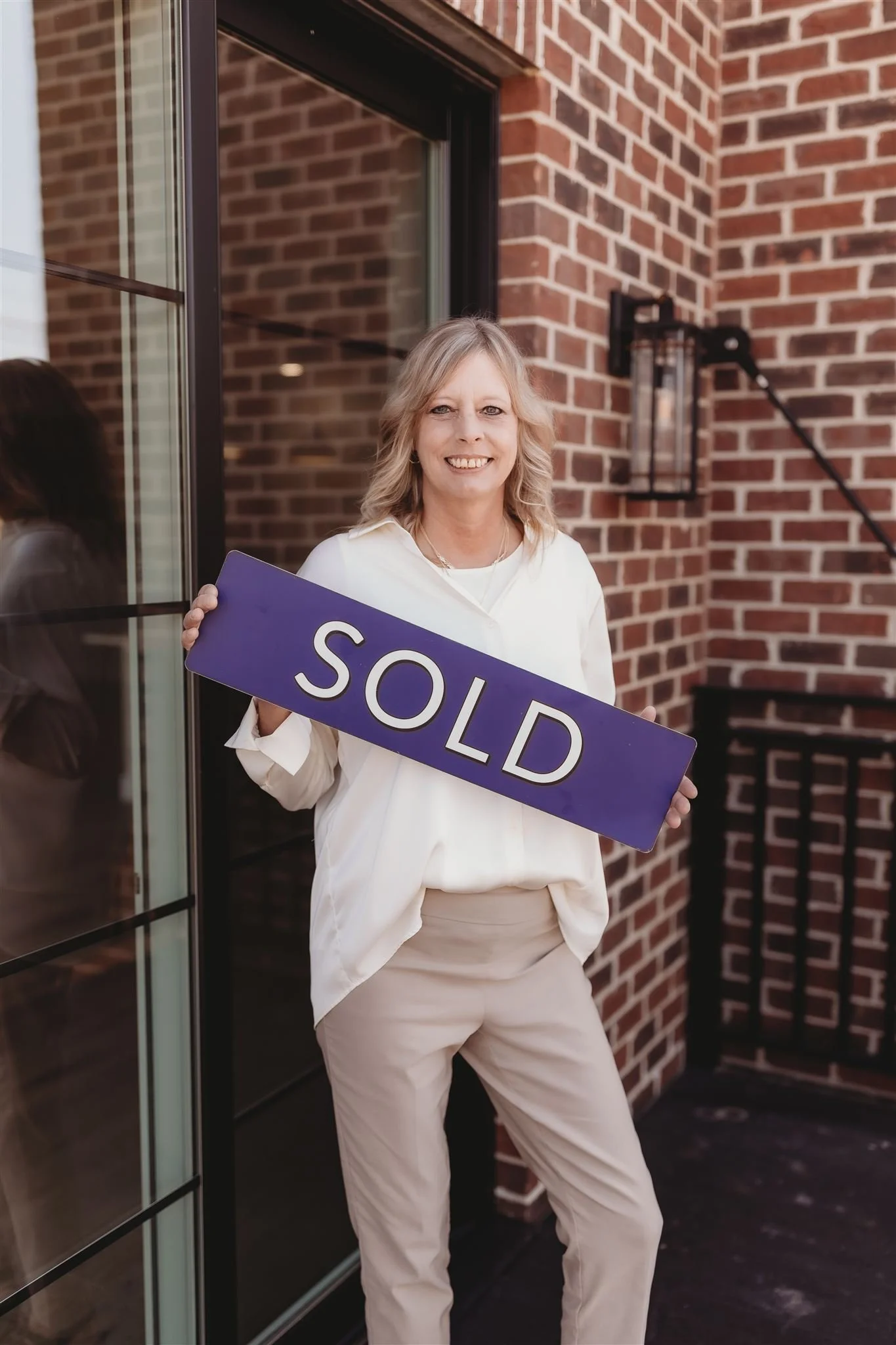 Woman smiling and holding a purple 'SOLD' sign outside a brick building.