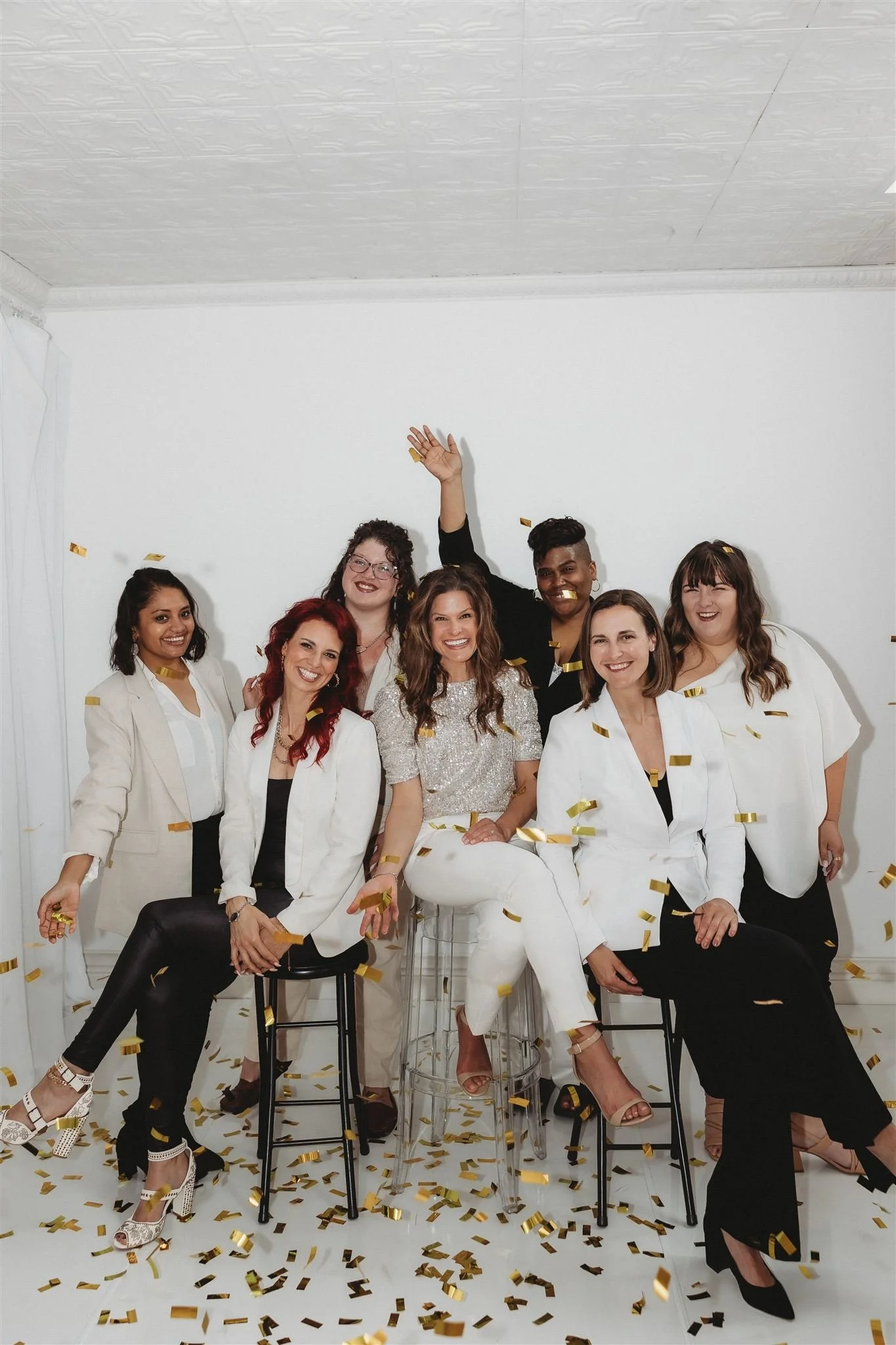 Group of seven diverse women celebrating with gold confetti, smiling, and sitting and standing in a plain white room.