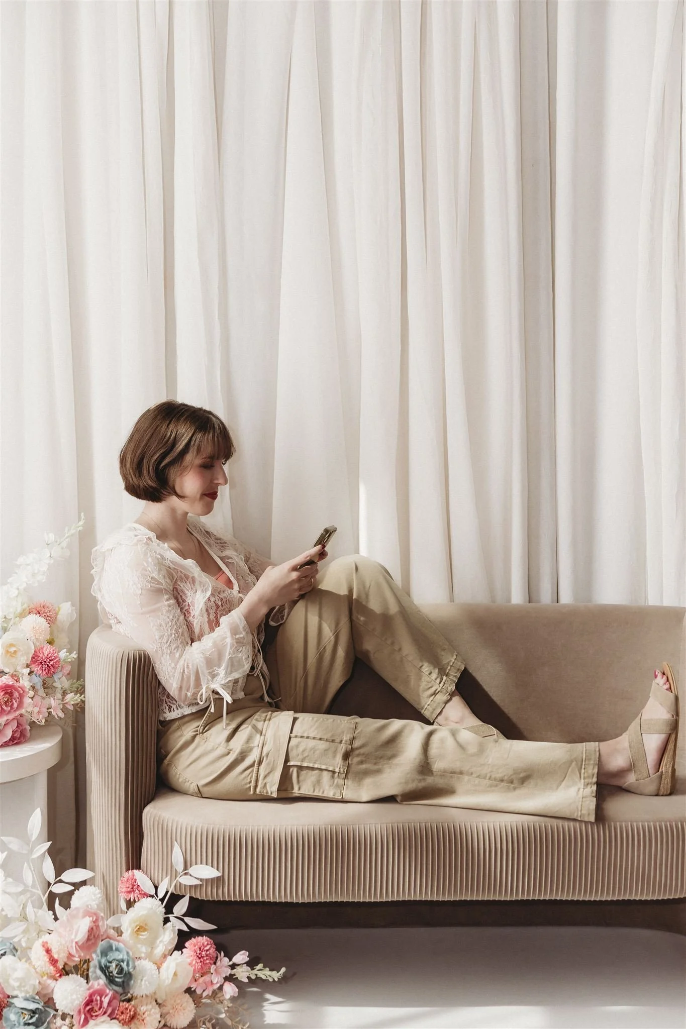 A woman with short brown hair, wearing beige pants and a lacy blouse, is sitting on a beige sofa, looking at her phone. She has a calm expression and is seated against a backdrop of white curtains, with a bouquet of pink, white, and blue flowers on a side table.