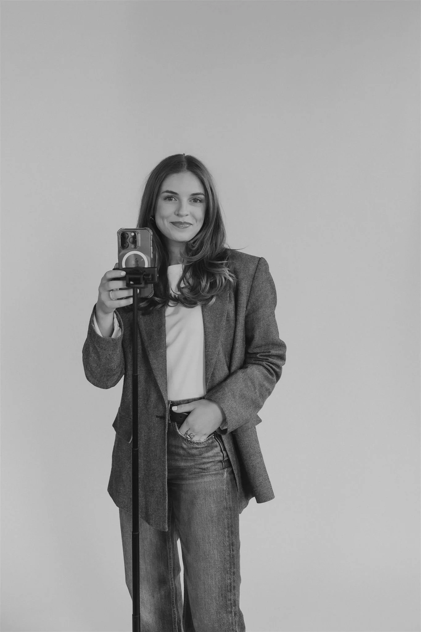 A woman takes a selfie in front of a plain wall, holding her phone with her left hand and smiling at the camera. She wears a blazer, a white top, and jeans.