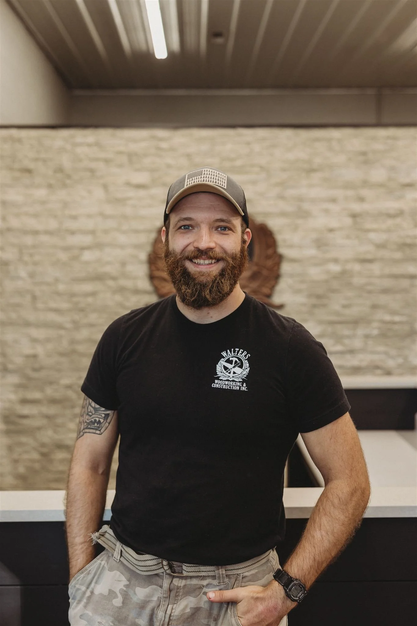 A man with a beard and a cap standing indoors in front of a beige brick wall, smiling and wearing a black T-shirt with a logo that reads 'WALTER'S WOODWORKING & CONSTRUCTION INC.', and camouflage shorts.